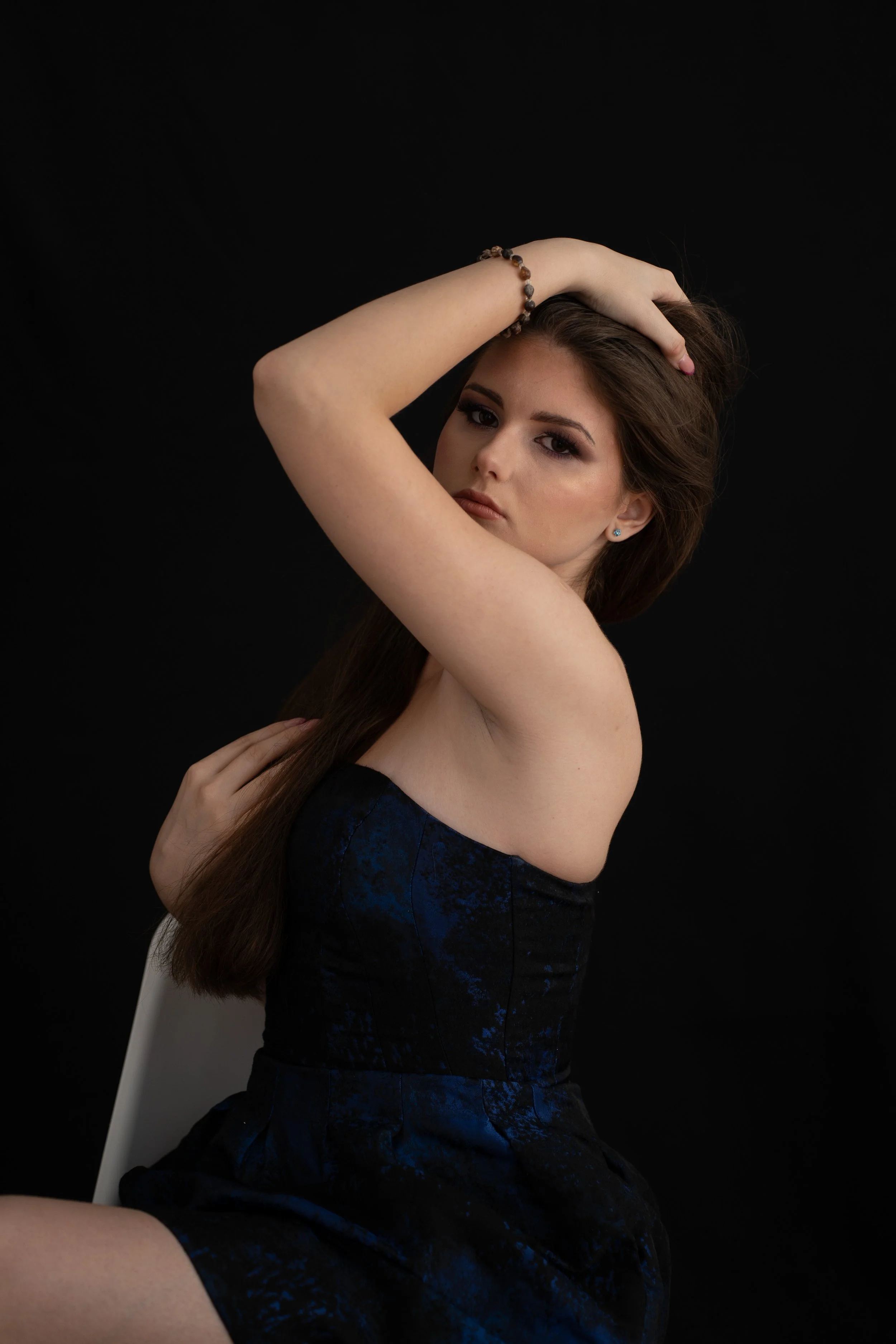 A woman with long brown hair, wearing earrings and a bracelet, posing with her left arm over her forehead and facing the camera against a black background.