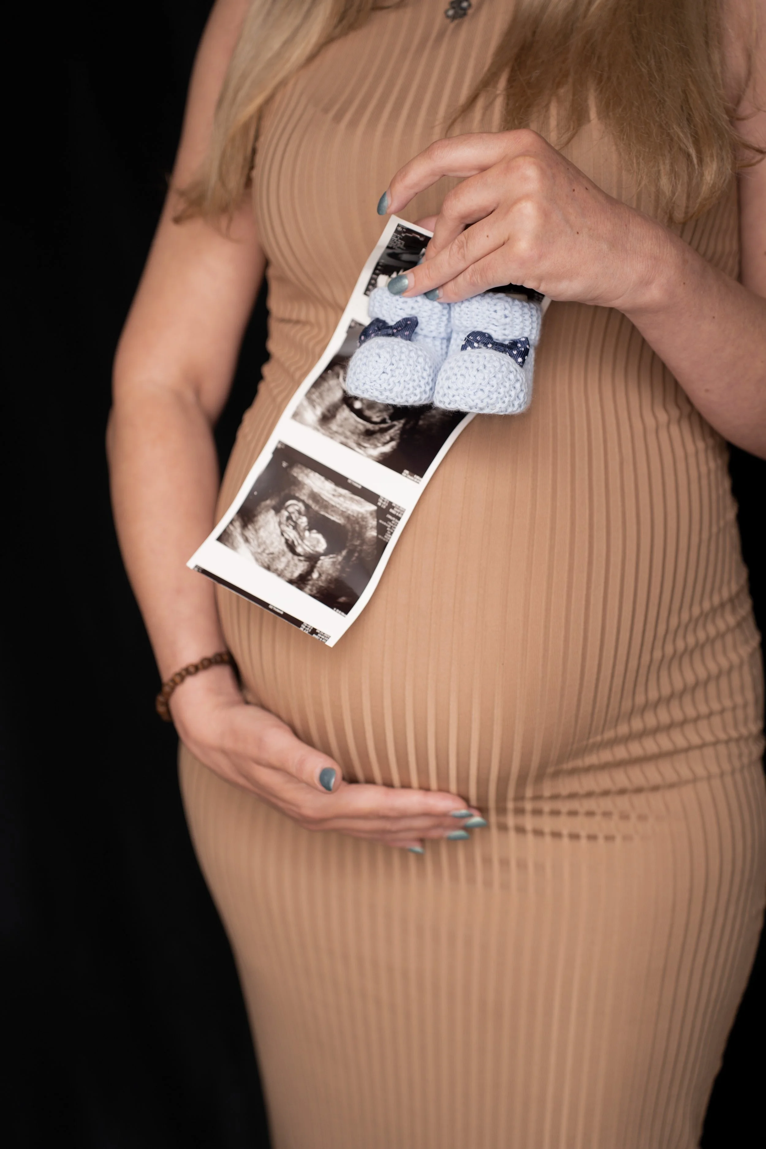 A pregnant woman holding ultrasound pictures against her baby bump, wearing a tan, ribbed, sleeveless dress, with her hand resting on her belly, and matching blue nail polish.