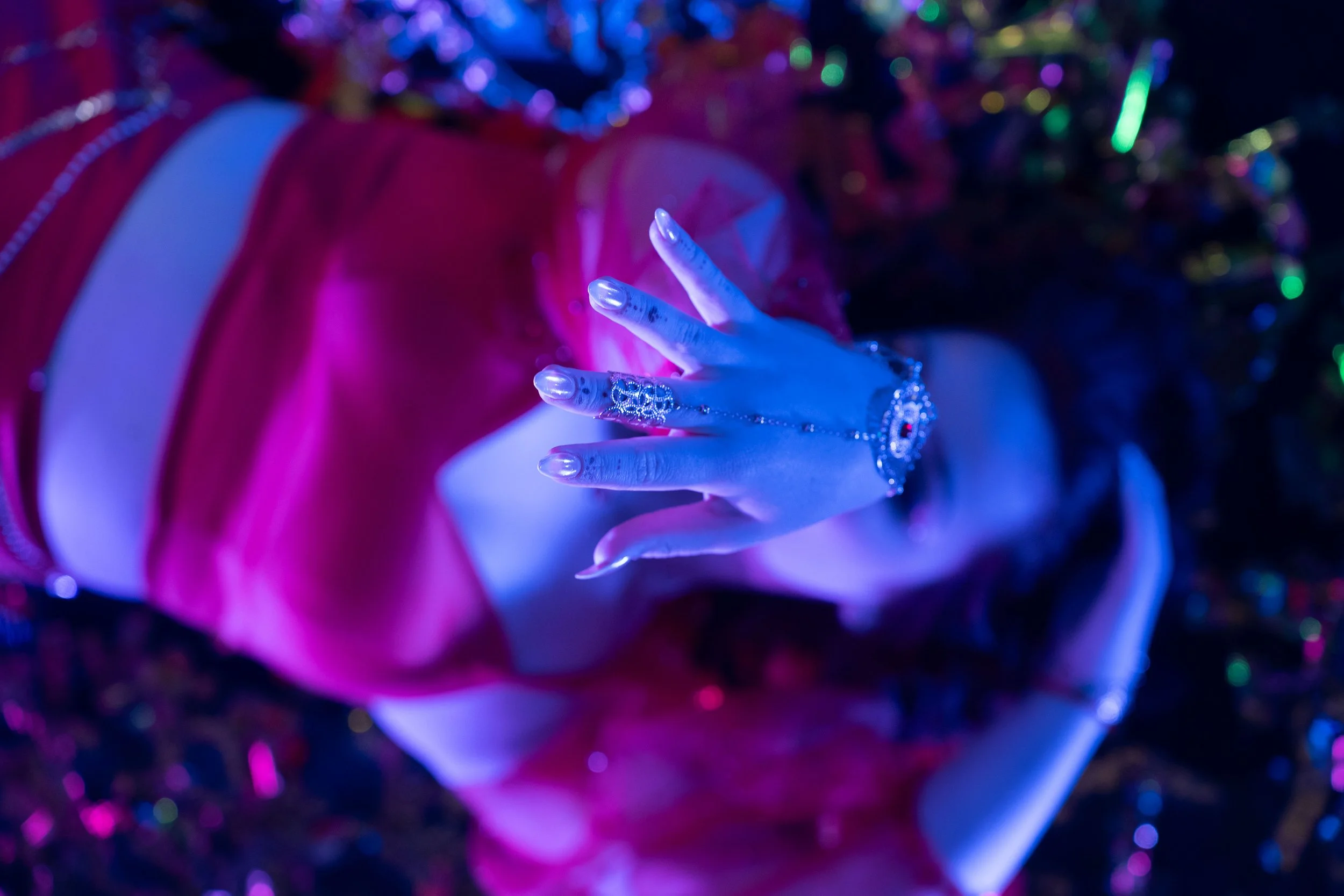 Close-up of a person's hand with jewelry, making a peace sign, illuminated in pink and blue lighting, with a blurred background of colorful lights.