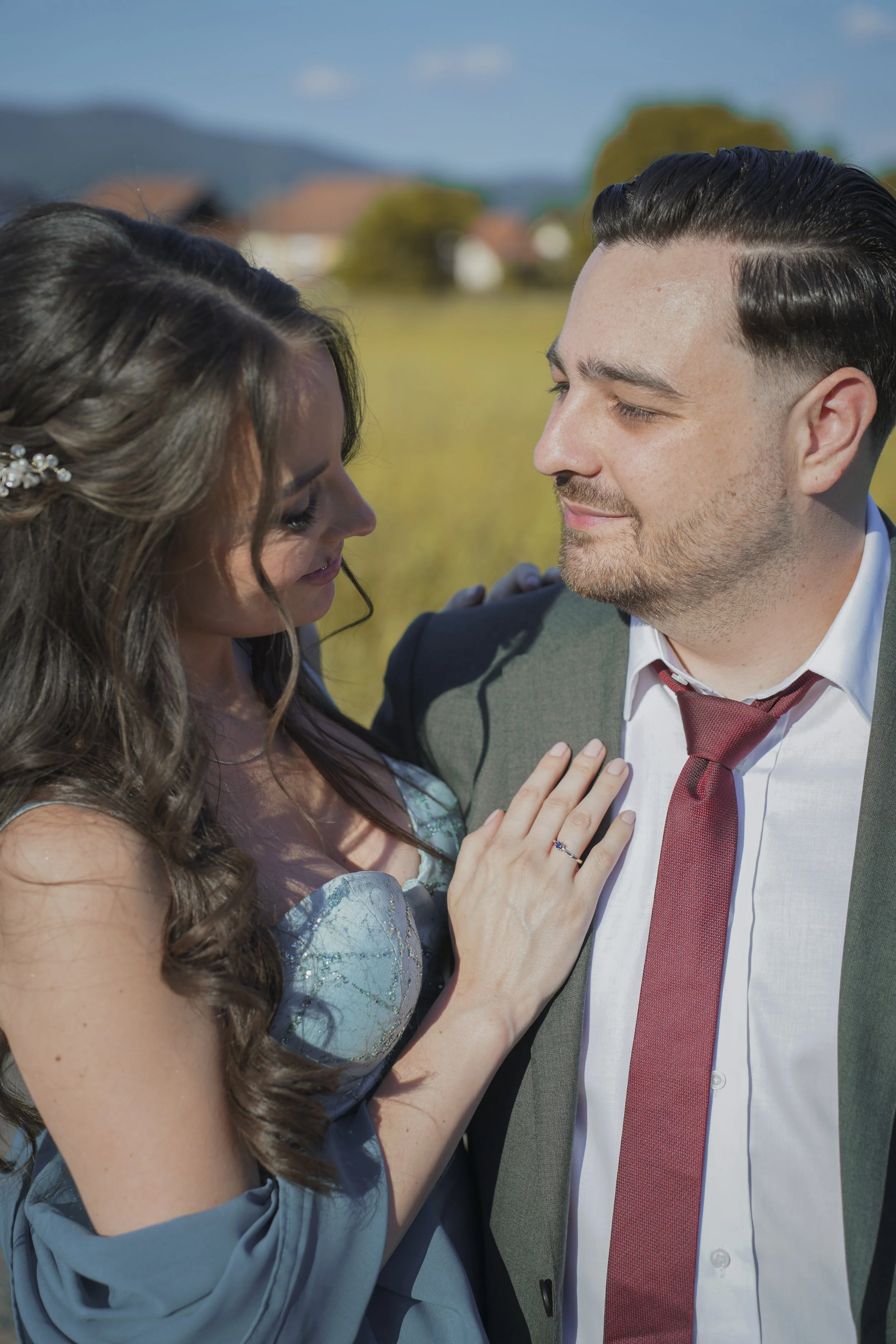 A couple, a woman with long wavy hair and a man with short dark slicked-back hair, dressed in formal attire, standing outdoors in a field with a blurred background of trees and houses, gazing into each other's eyes intimacy, touching.