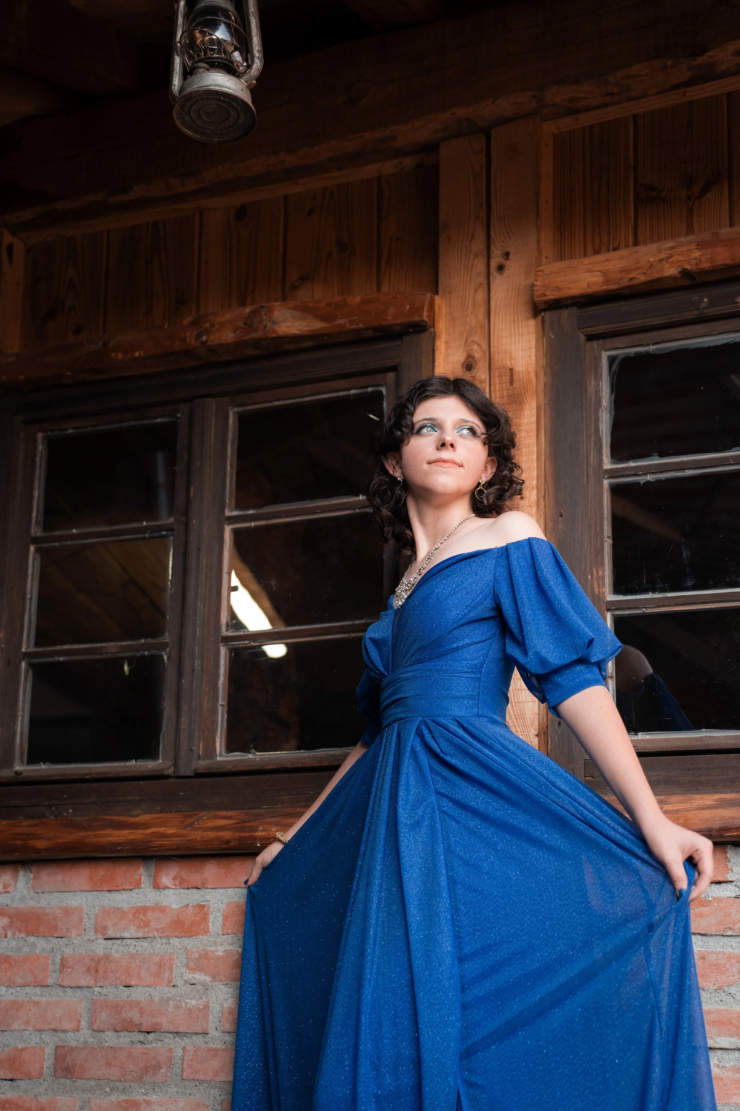 A woman in a blue dress standing outside in front of a wooden wall with windows, with her face turned upward.