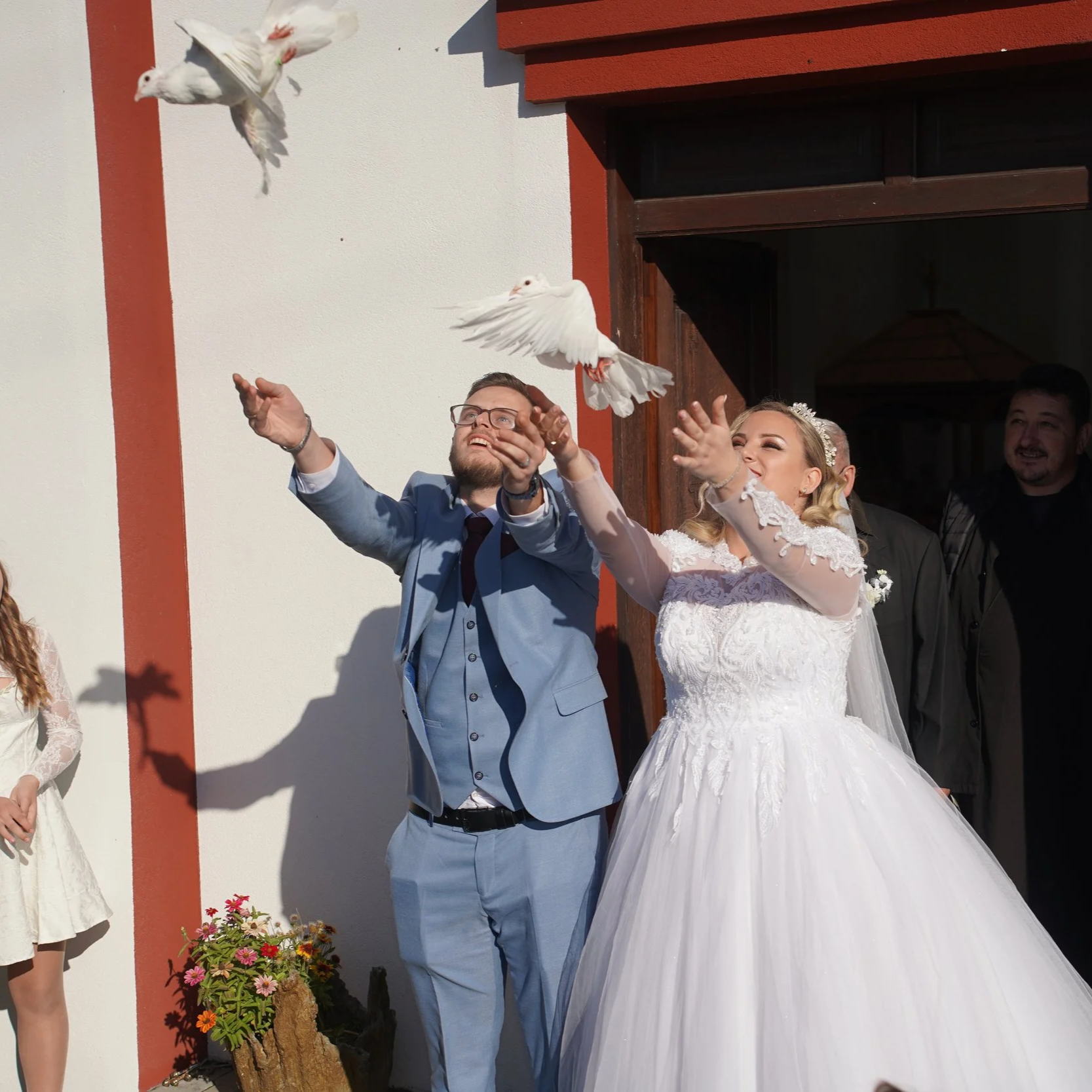 A groom and bride release white doves during their wedding ceremony outside a building with a white and red wall. The groom is wearing a light blue suit and glasses, and the bride is in a white wedding dress with lace details and a tiara.