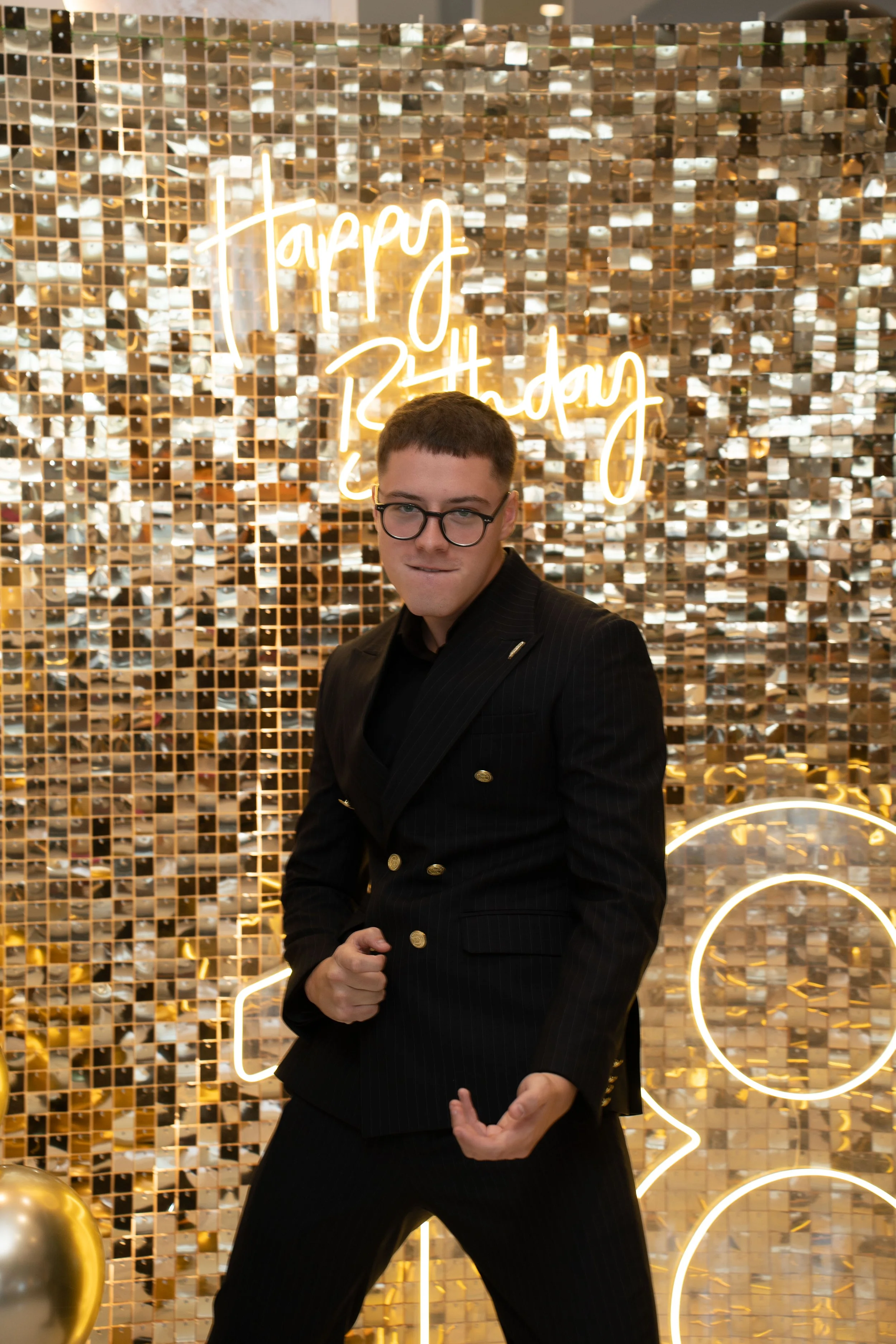 Young man wearing glasses and a dark tuxedo posing in front of a shiny gold mosaic wall with neon sign that reads 'Happy Birthday'.