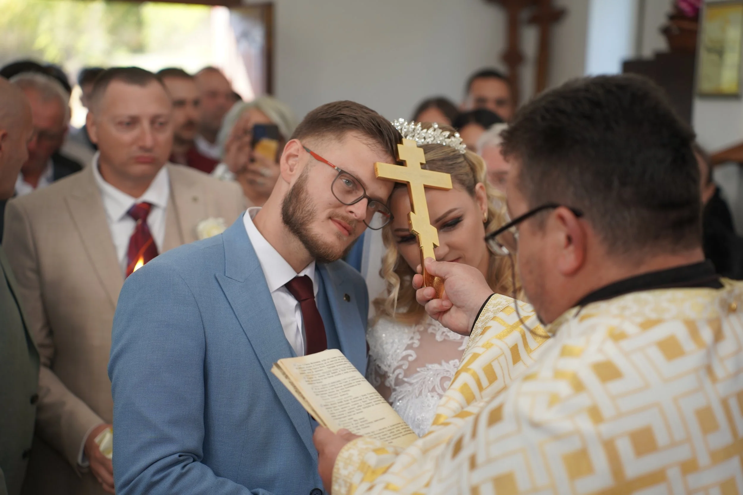 A wedding ceremony with a groom and bride kneeling before a priest, holding a cross during their vows, while guests look on.