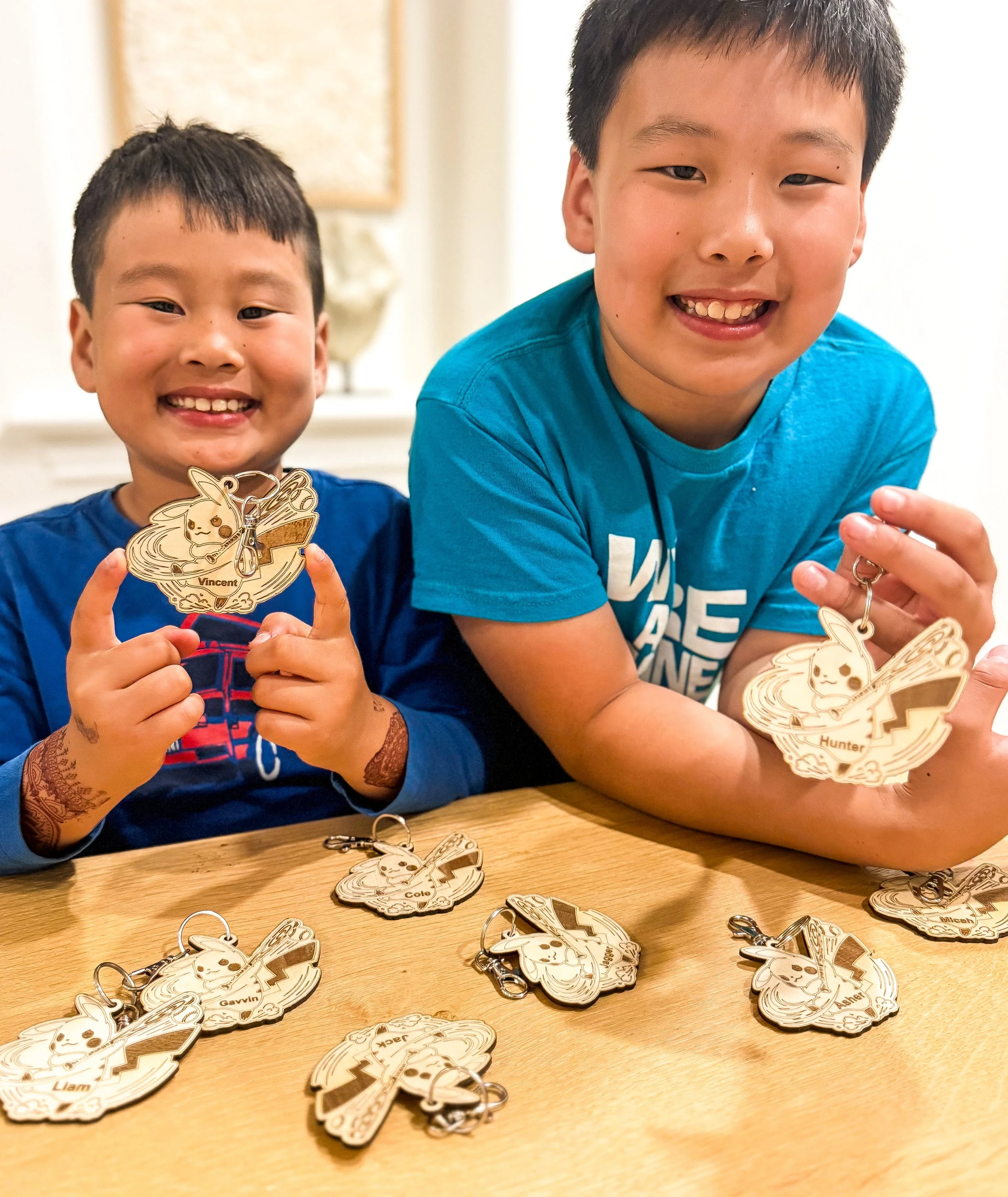 Two smiling boys showing Pokémon-themed keychains on a wooden table, with more keychains scattered on the table, in a home setting.