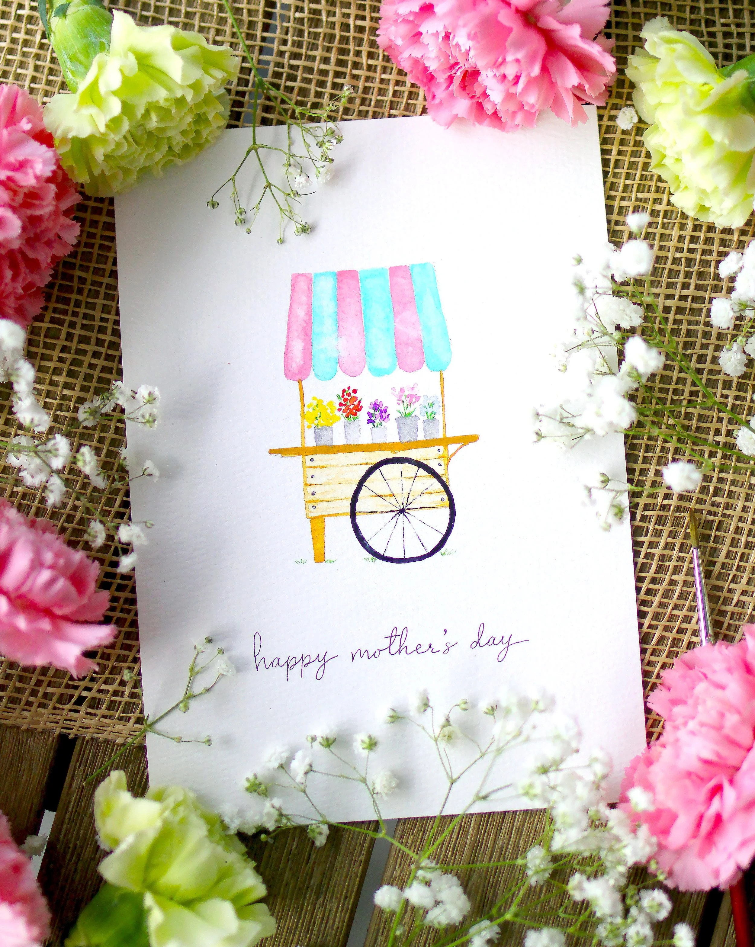 Handmade Mother's Day card with a colorful flower cart illustration and the message 'happy mother's day' surrounded by pink and white flowers on a woven background.