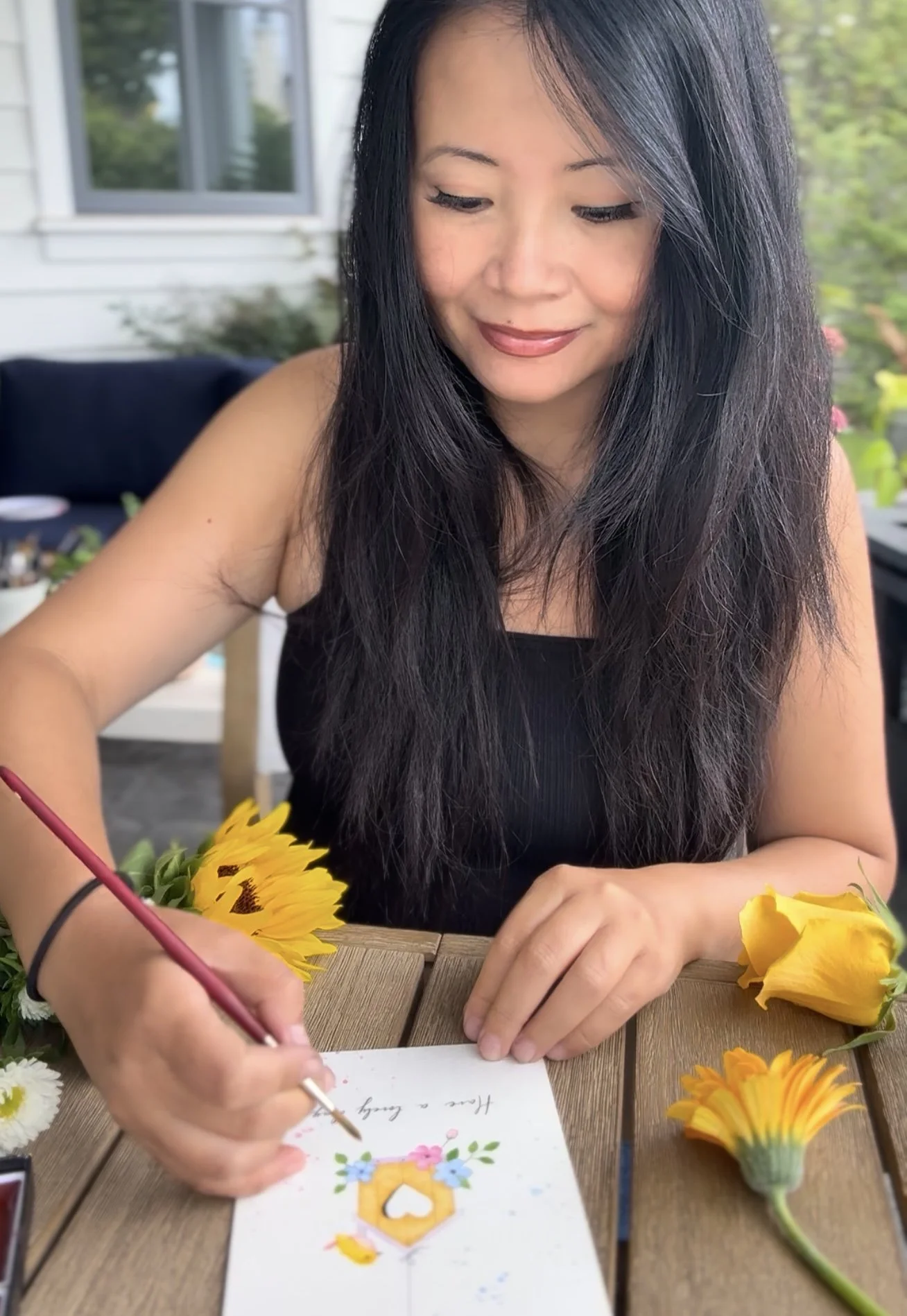 Woman with long black hair writing on a greeting card with flowers on the table, surrounded by yellow and orange flowers, on a wooden table outside with a house and greenery in the background.