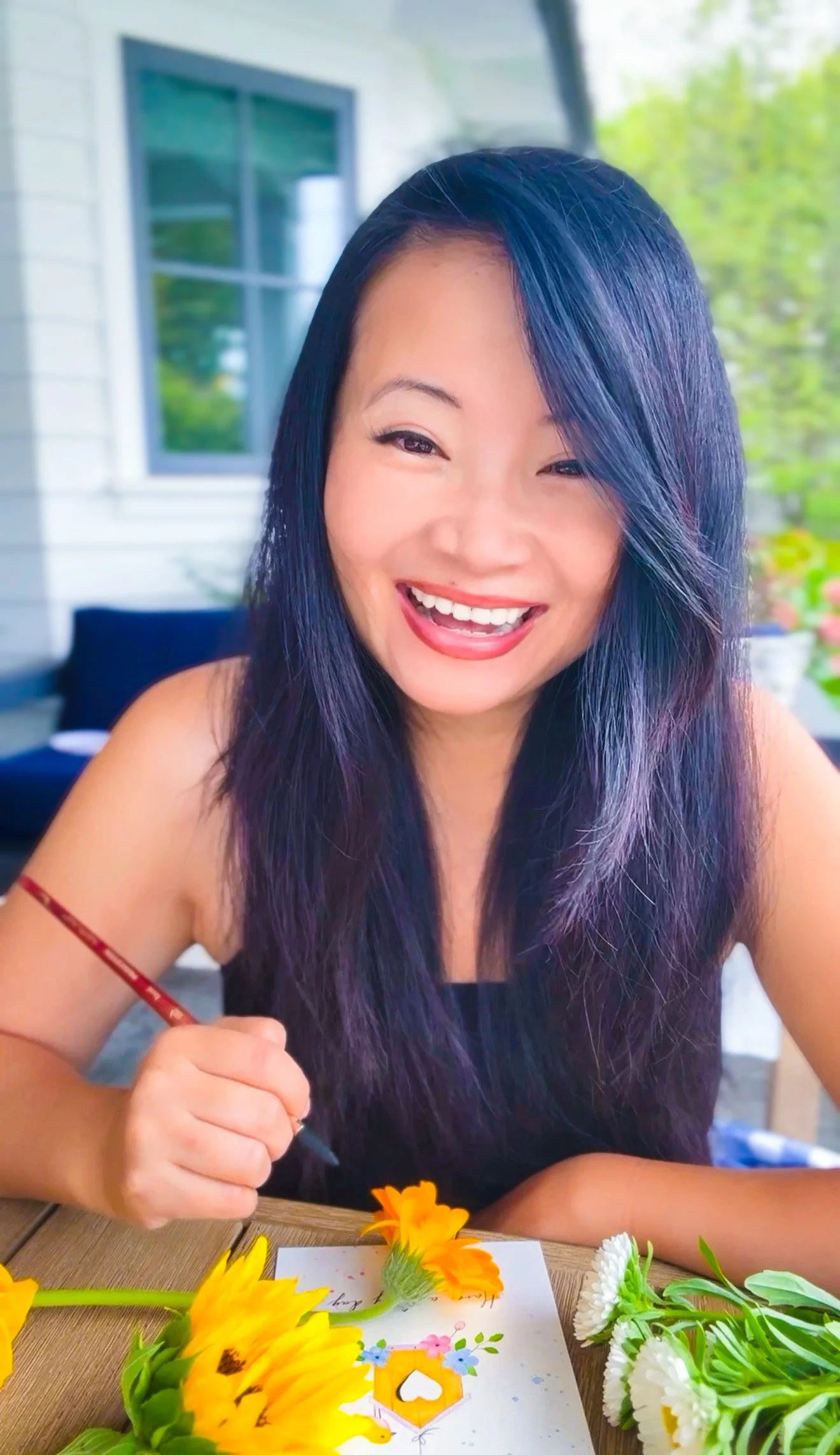 A woman with long purple hair is smiling while painting a colorful picture with flowers and a birdhouse on paper. There are various flowers on the table.