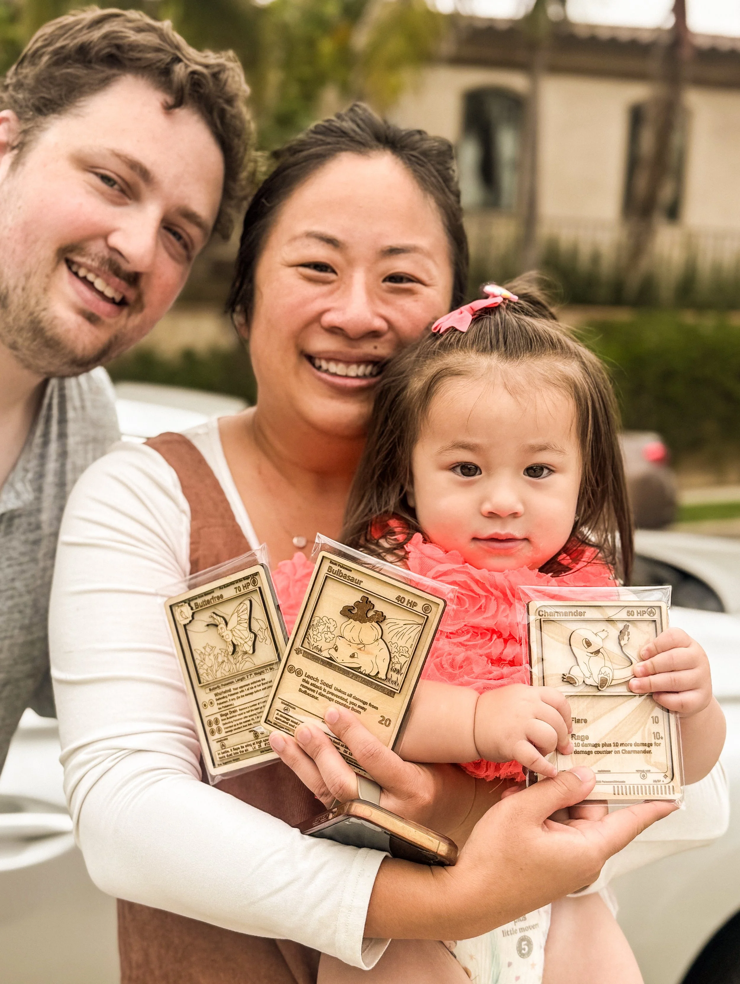 A family of three, including a man, a woman, and a young girl, posing outdoors with the girl holding three Pokémon cards.