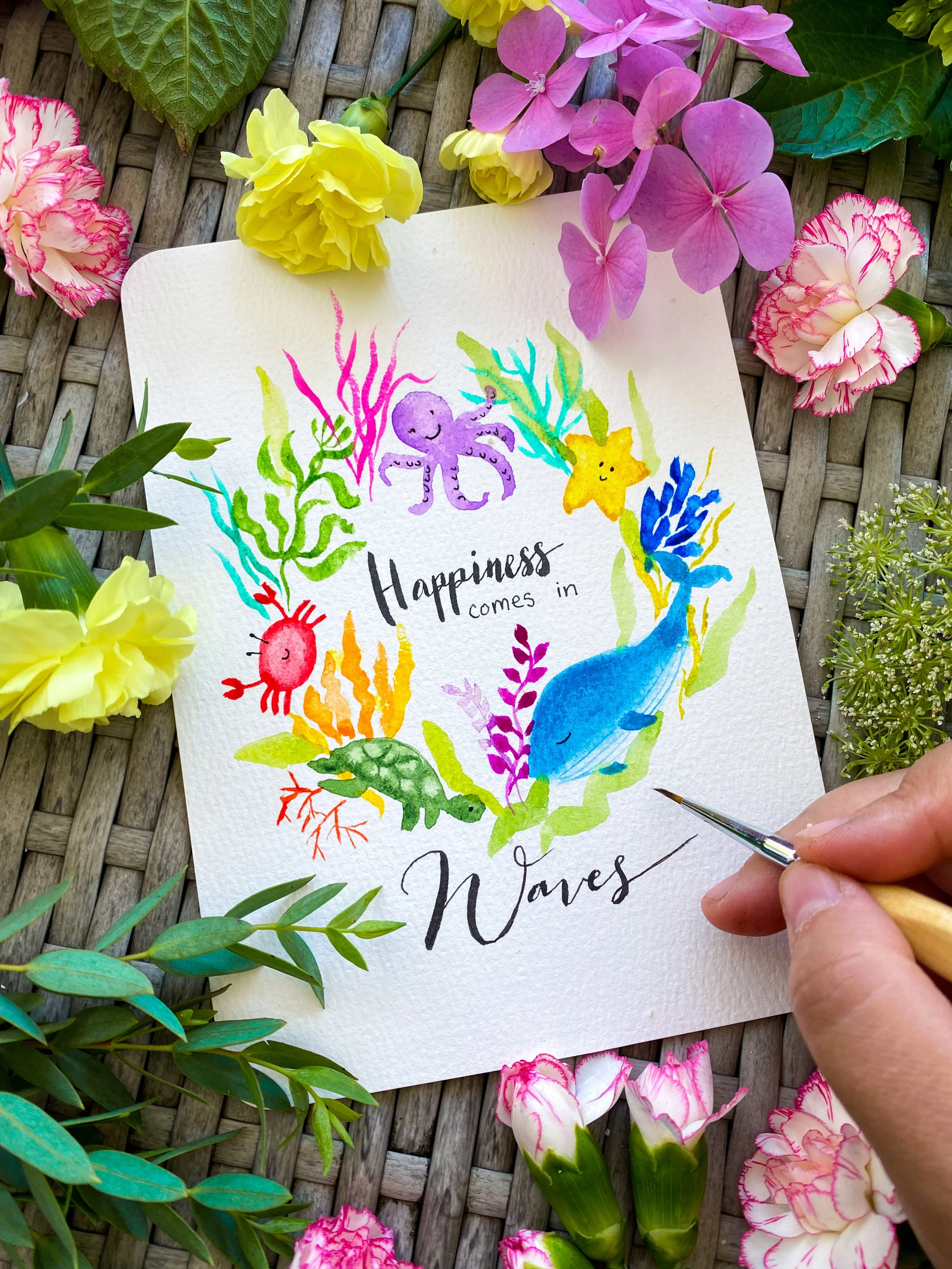 Colorful watercolor illustration of underwater sea creatures such as an octopus, starfish, whale, turtle, crab, and various coral and seaweed, with the phrase "Happiness comes in waves" written in black calligraphy, surrounded by pink, yellow, purple, and green flowers and foliage.
