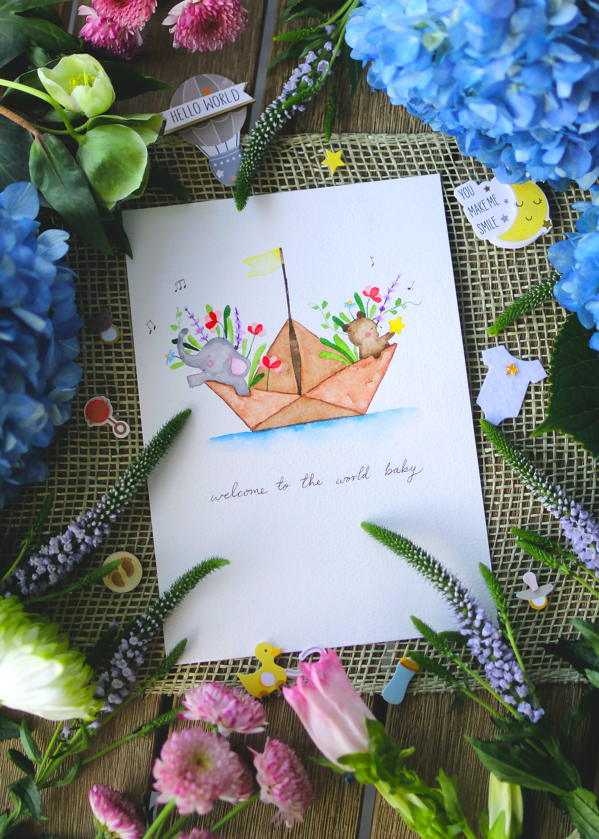 A colorful watercolor illustration of a paper boat with tiny animals and flowers inside. The drawing includes a gray elephant and a brown bear, surrounded by pink, purple, and red flowers, with the phrase 'welcome to the world baby' written underneat