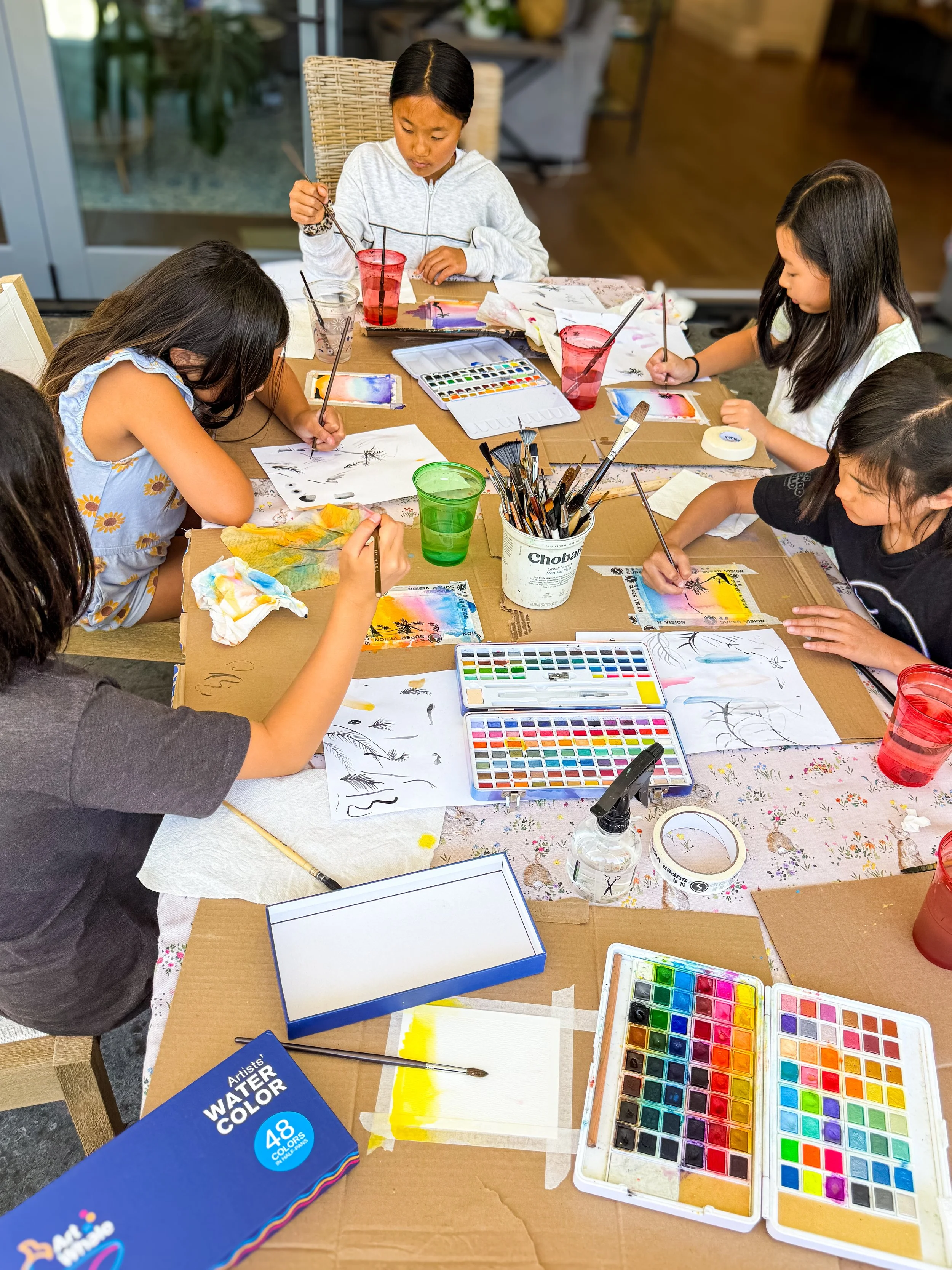 Group of children painting watercolor pictures at a table with art supplies and water cups.
