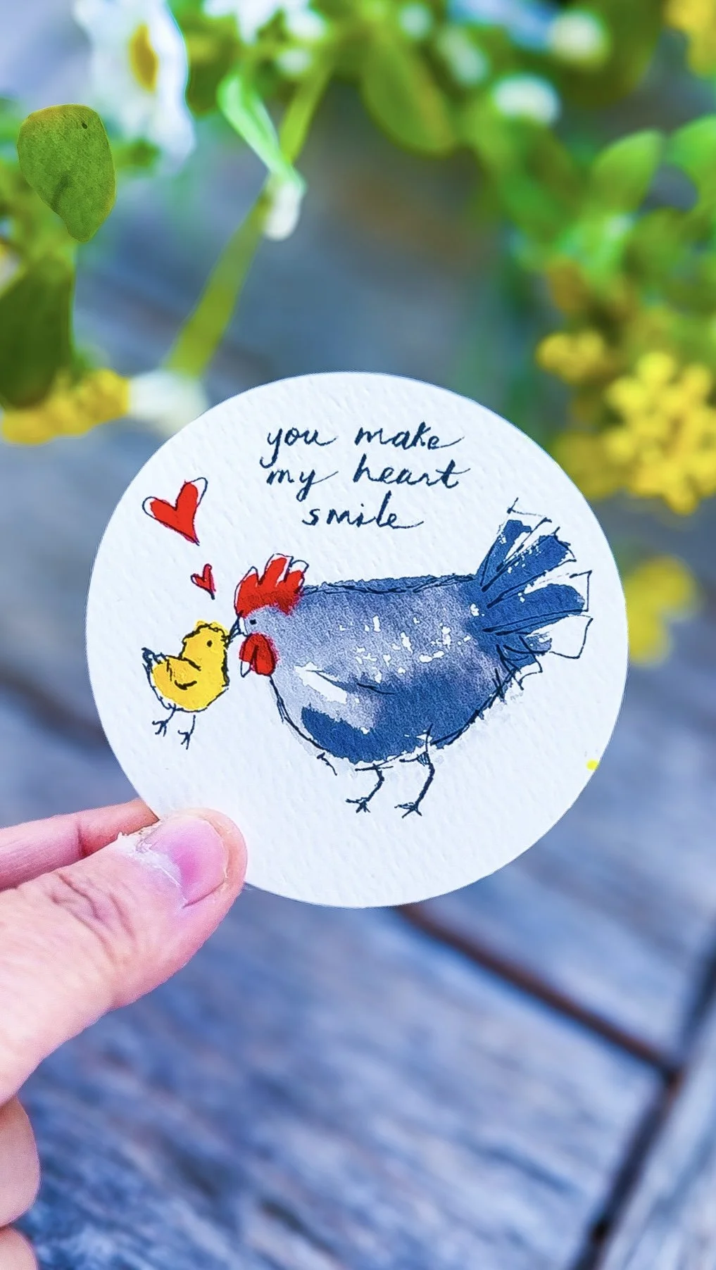 A circular paper with a watercolor illustration of a hen and a chick, accompanied by handwritten text and red hearts, held by a person's hand outdoors.