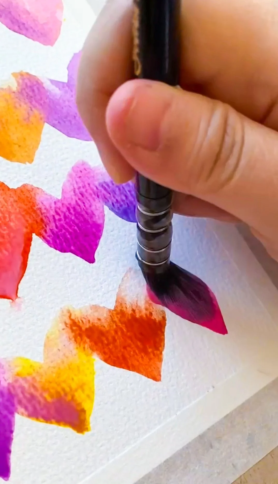 Close-up of a hand holding a black paintbrush, painting colorful heart shapes on a white canvas.