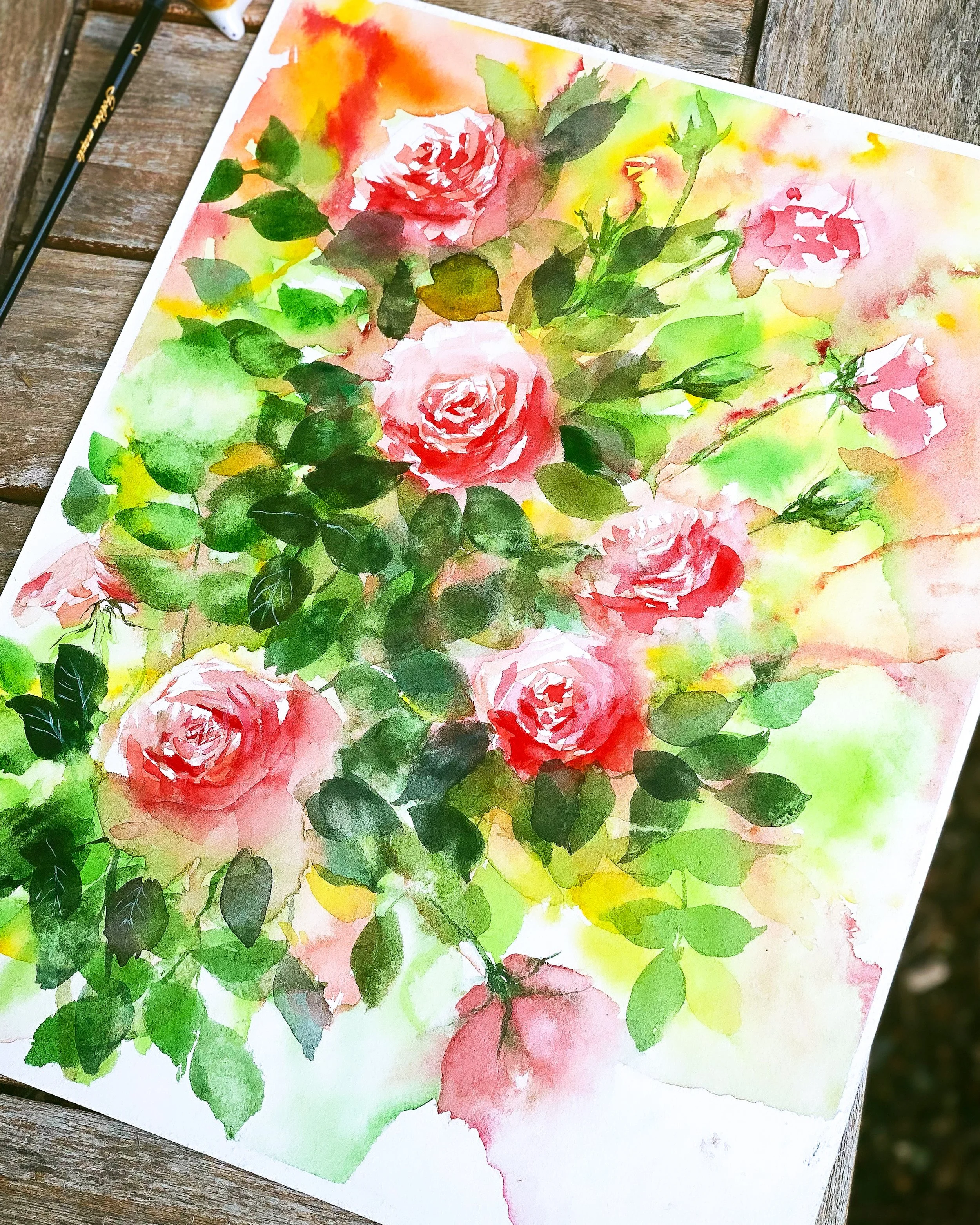 Watercolor painting of pink roses with green foliage on paper, placed on a wooden surface.