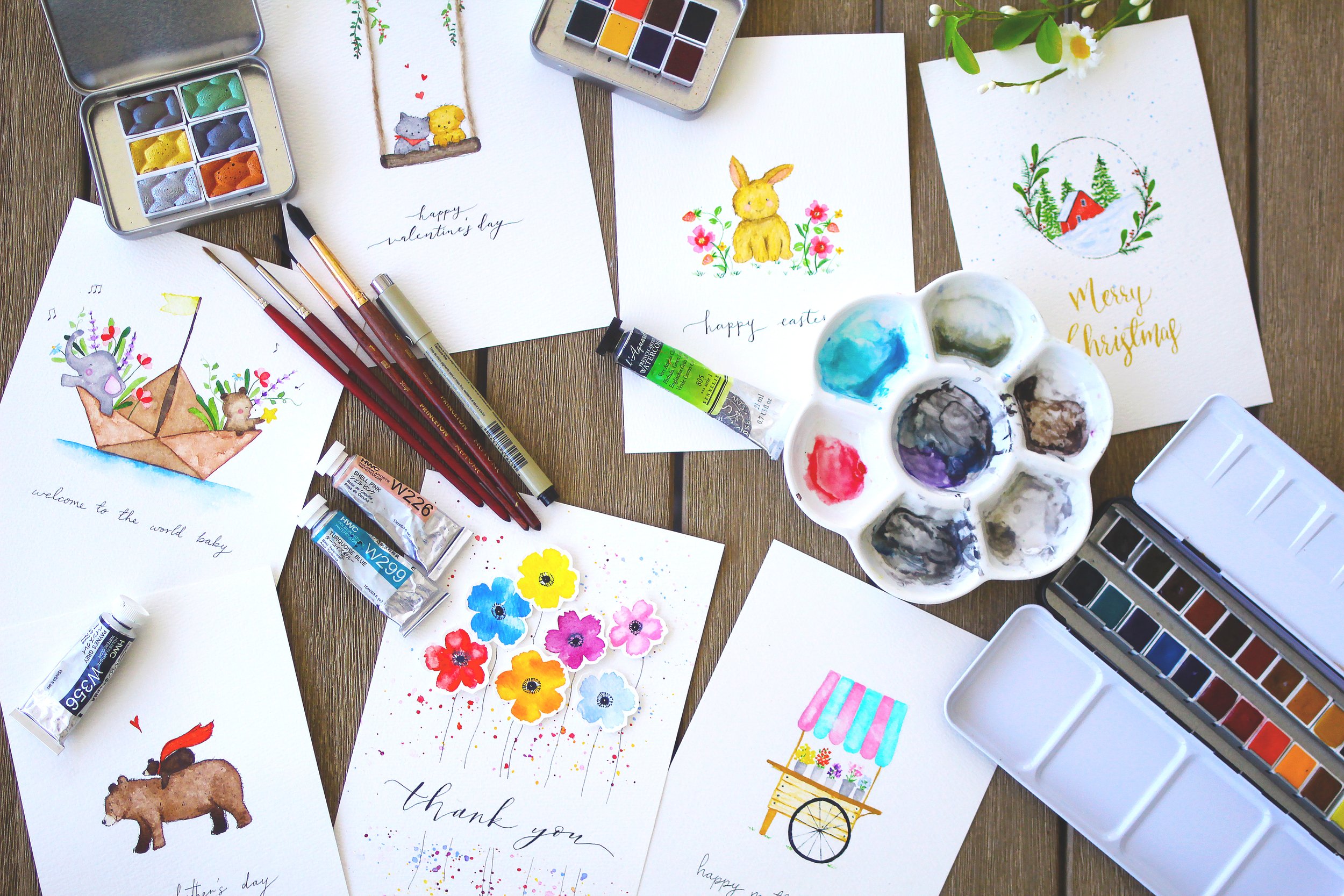 A table with watercolor paintings and greeting cards for various occasions, watercolor paints, brushes, and a palette on a wooden surface.