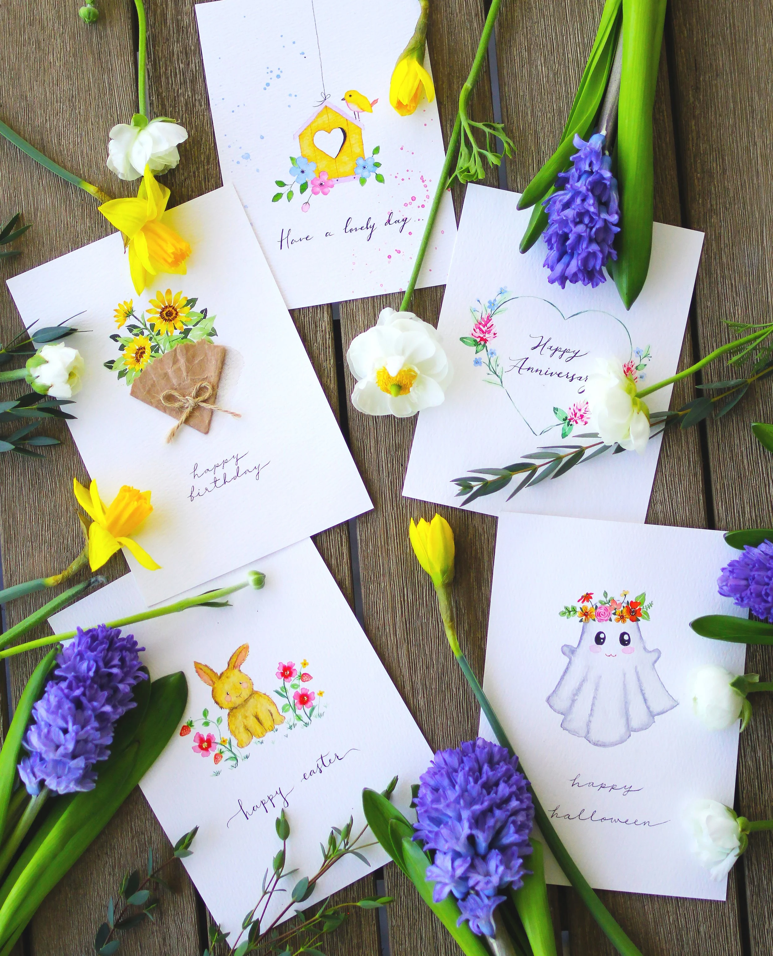 Collection of handmade greeting cards with floral and cute animal illustrations, surrounded by colorful spring flowers including daffodils, hyacinths, and tulips, on a wooden surface.