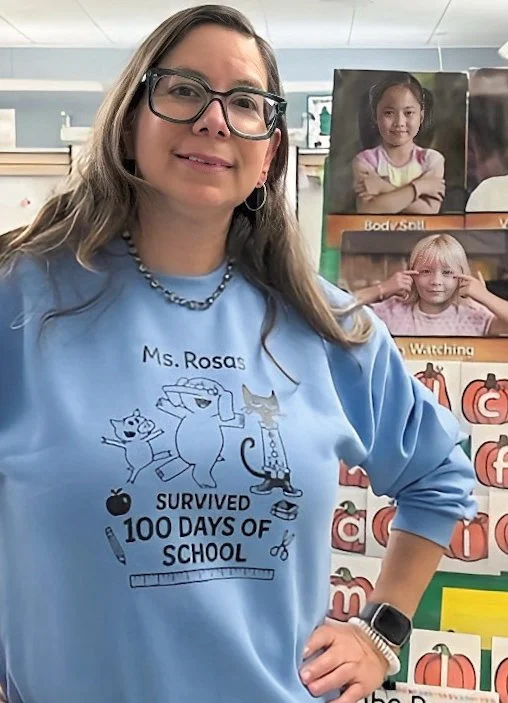 Teacher smiling in classroom, wearing a blue sweatshirt that says 'Ms. Rosa's Survived 100 Days of School', with educational posters and children's photos in the background.