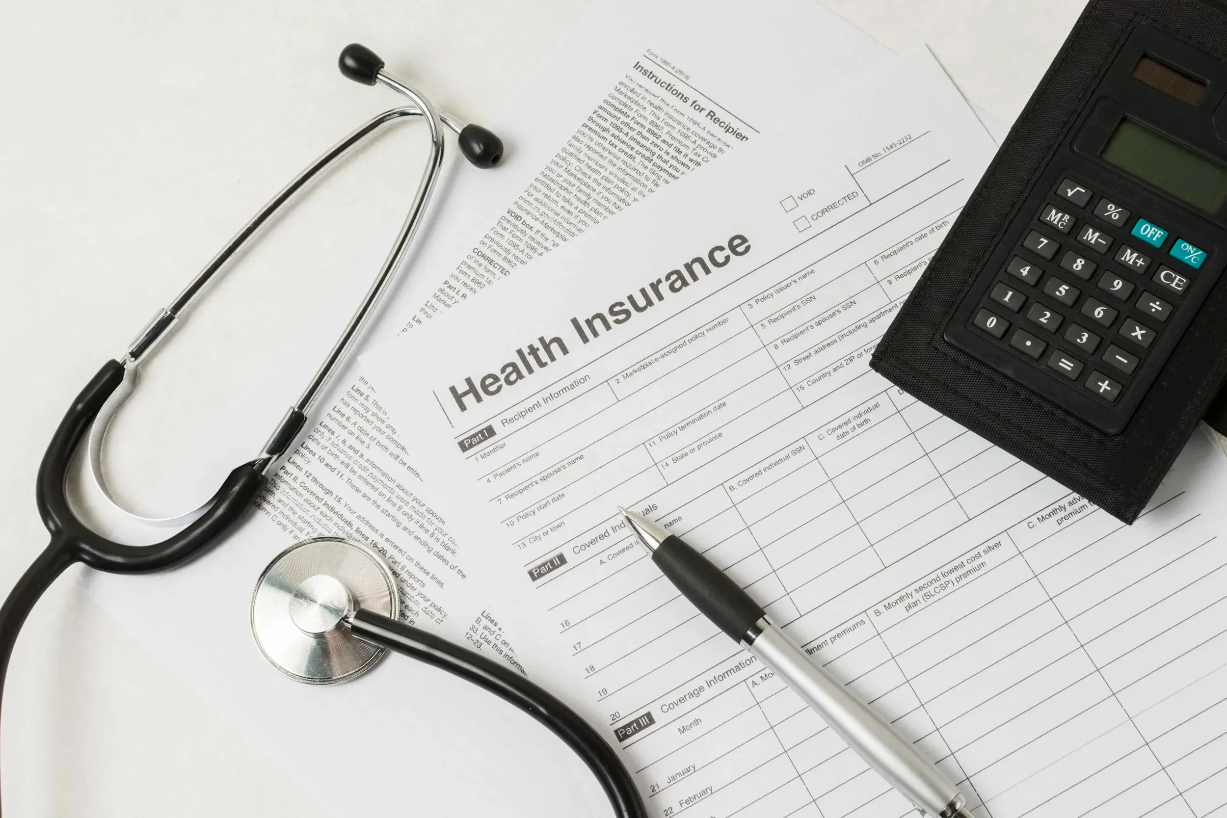 A stethoscope, health insurance form, pen, calculator, and some papers on a white surface.
