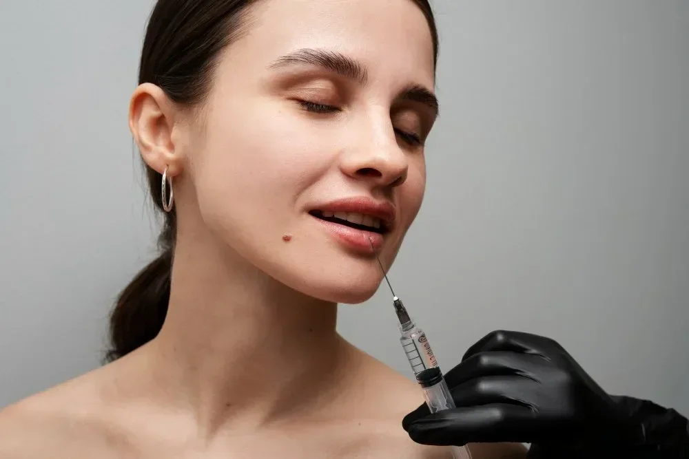 A woman with closed eyes receives a lip injection from a medical professional wearing black gloves.