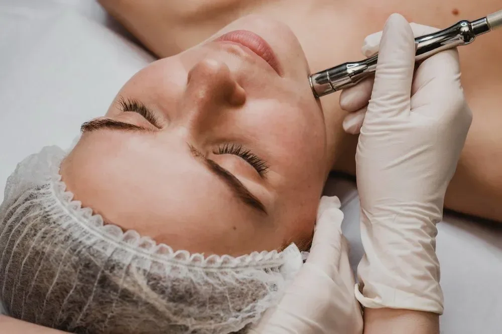 A person receiving a facial treatment with a beauty device, lying on a bed with a hairnet, while a technician in gloves operates the device.