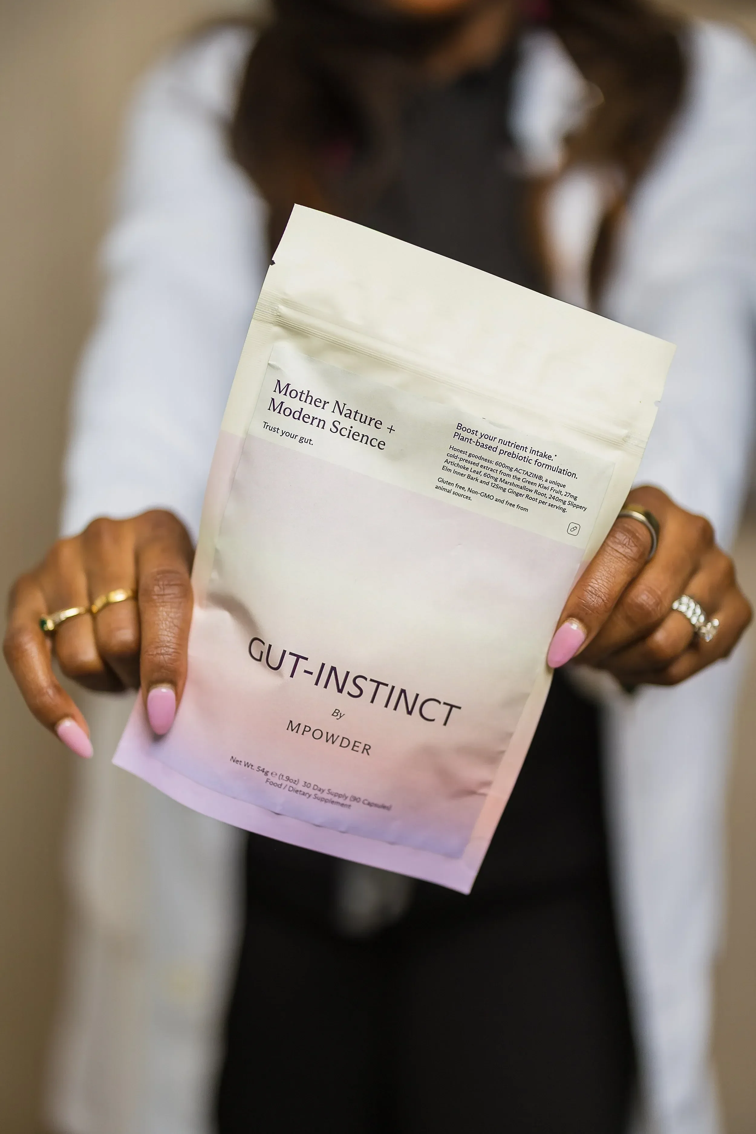A woman with light pink nails holding a white packet labeled "Gut-Instruct" and part of her outfit is visible, including rings on her fingers, a black top, and a white shirt. The background is blurred.
