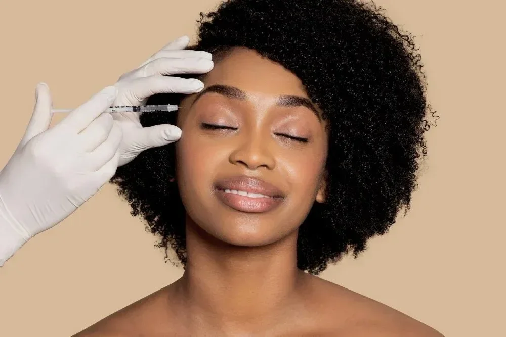 A woman relaxing with eyes closed while a healthcare professional prepares to give her an injection in her forehead.