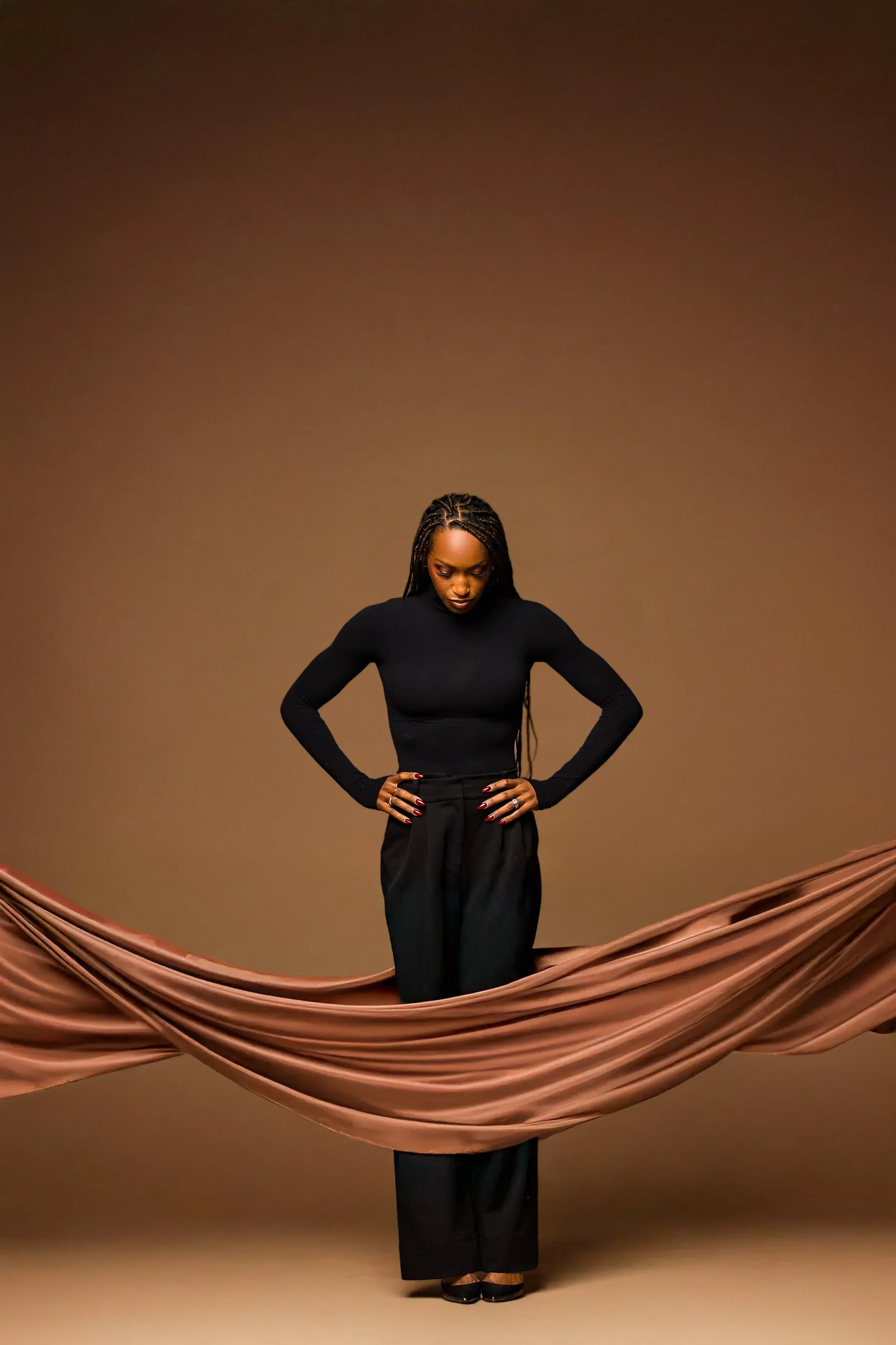 Dr. Spence wearing a black long-sleeve top and black pants, standing with her hands on her hips in front of a plain brown background, with a large satin fabric draped in the foreground.