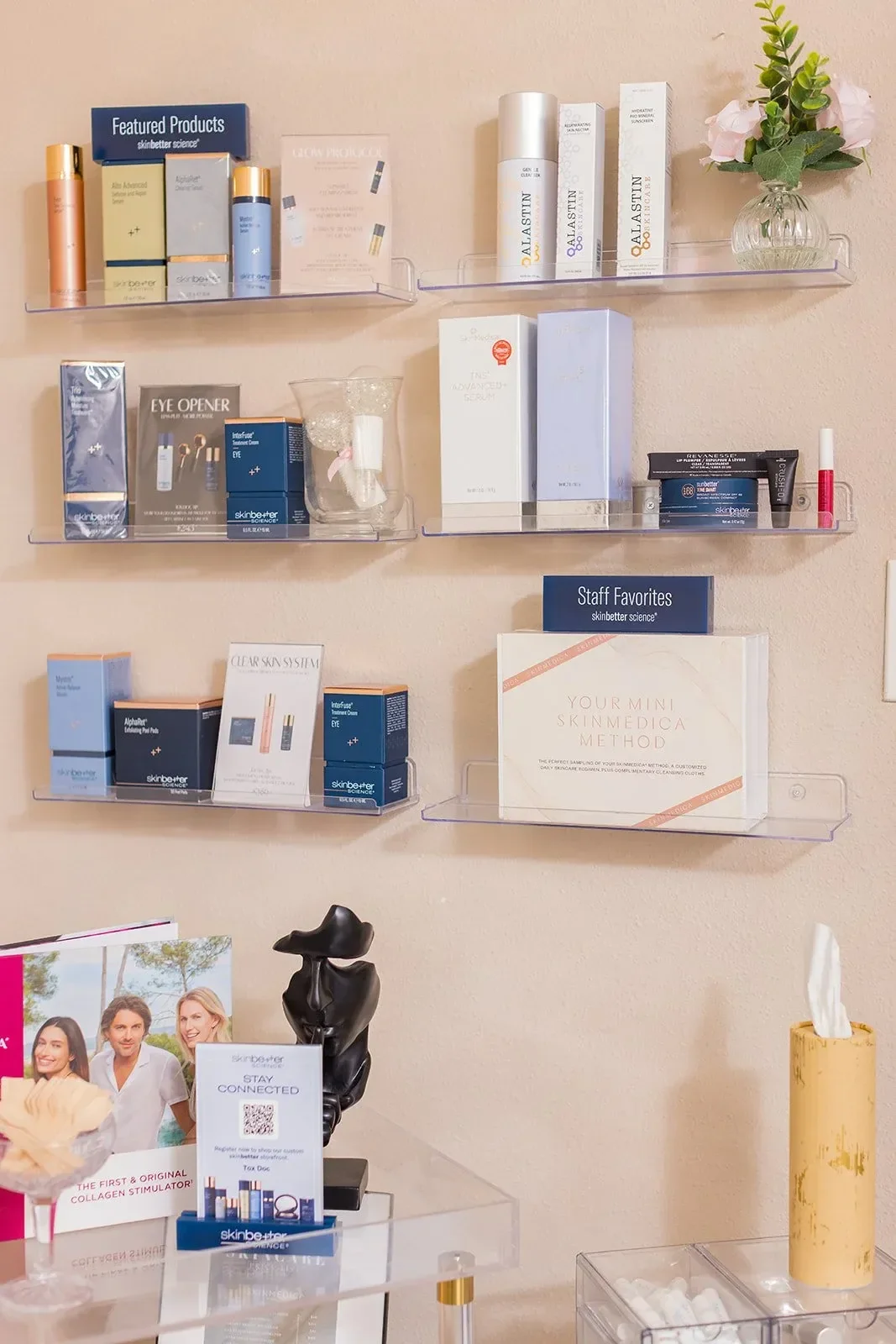 Display of skincare and beauty products on clear wall shelves with a small flower arrangement, a black sculpture, and promotional materials in the background.