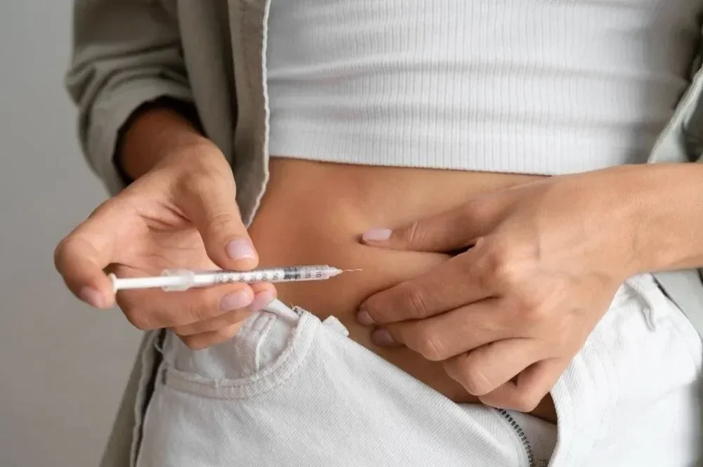 A person holding a syringe with a needle, preparing to administer an injection into their abdomen. The person is wearing a white crop top and light-colored pants.