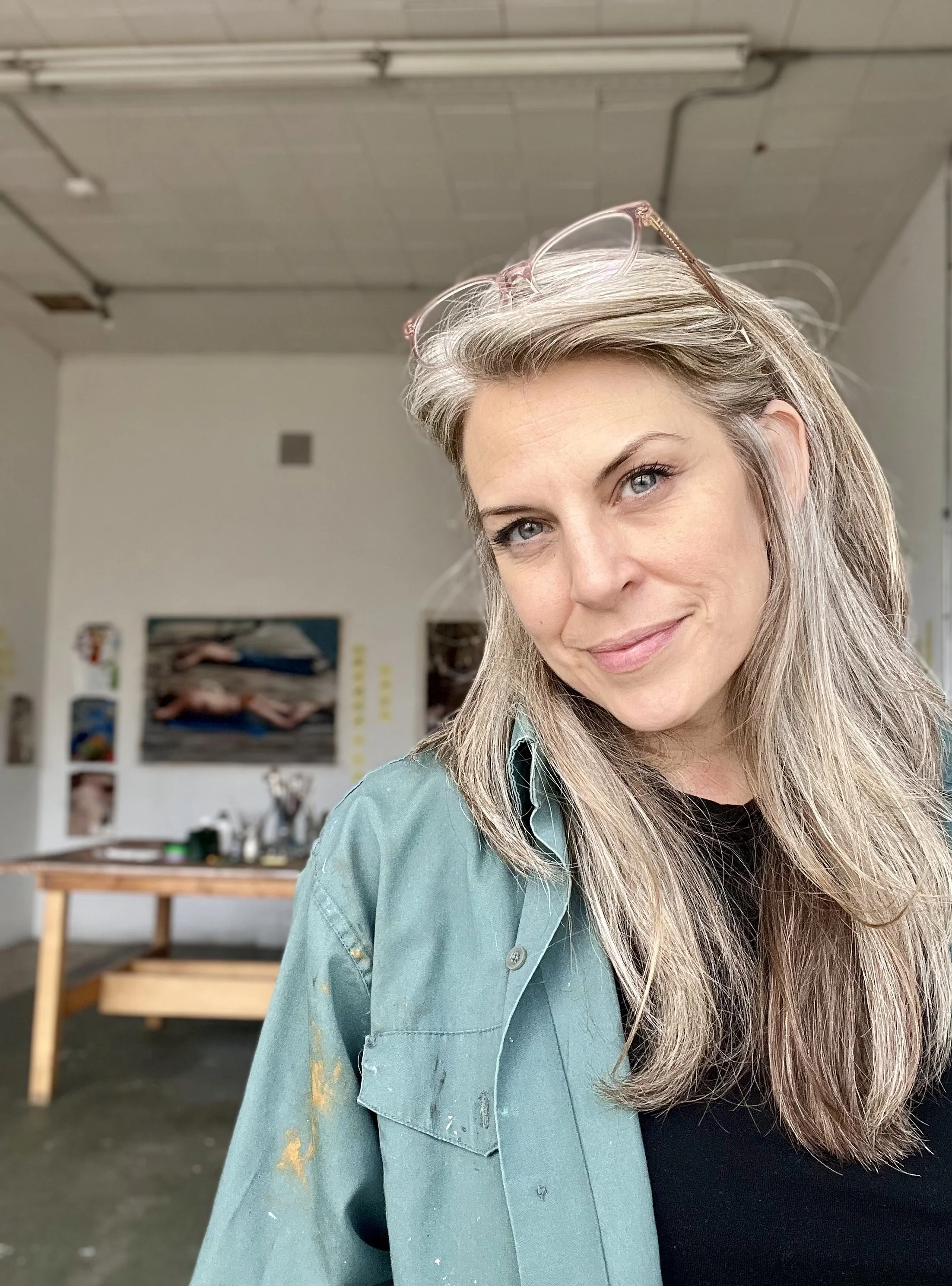 A woman with gray hair and blue eyes smiling in an art studio. She is wearing glasses on her head and a green shirt with paint stains.