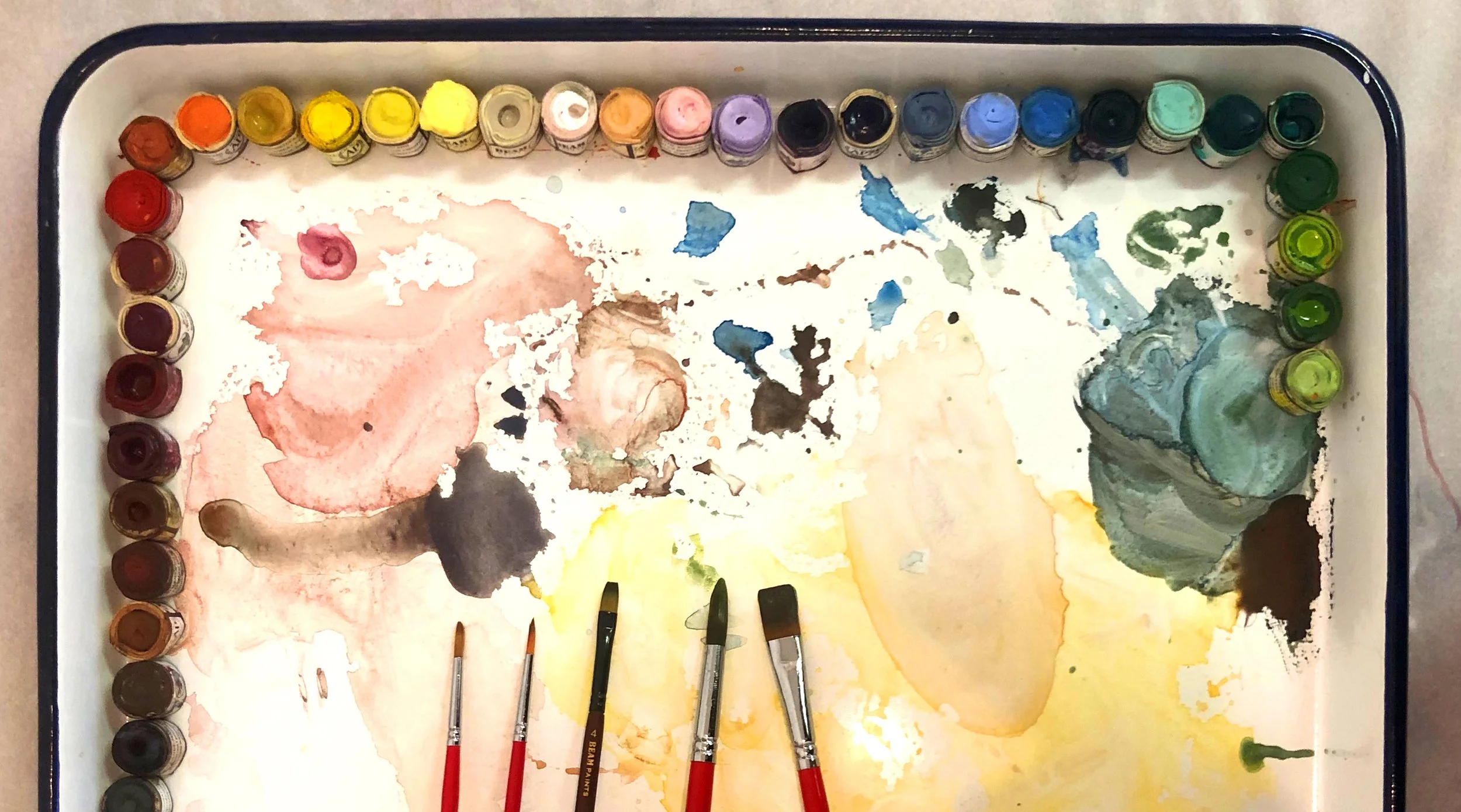Watercolor paint set with various colors on a tray and five paintbrushes.