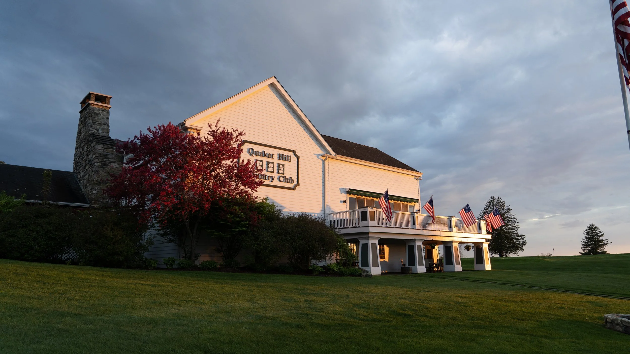 White building labeled 'Quaker Hill Country Club' with a porch decorated with American flags, surrounded by trees with red and green foliage, under a cloudy sky at sunset.
