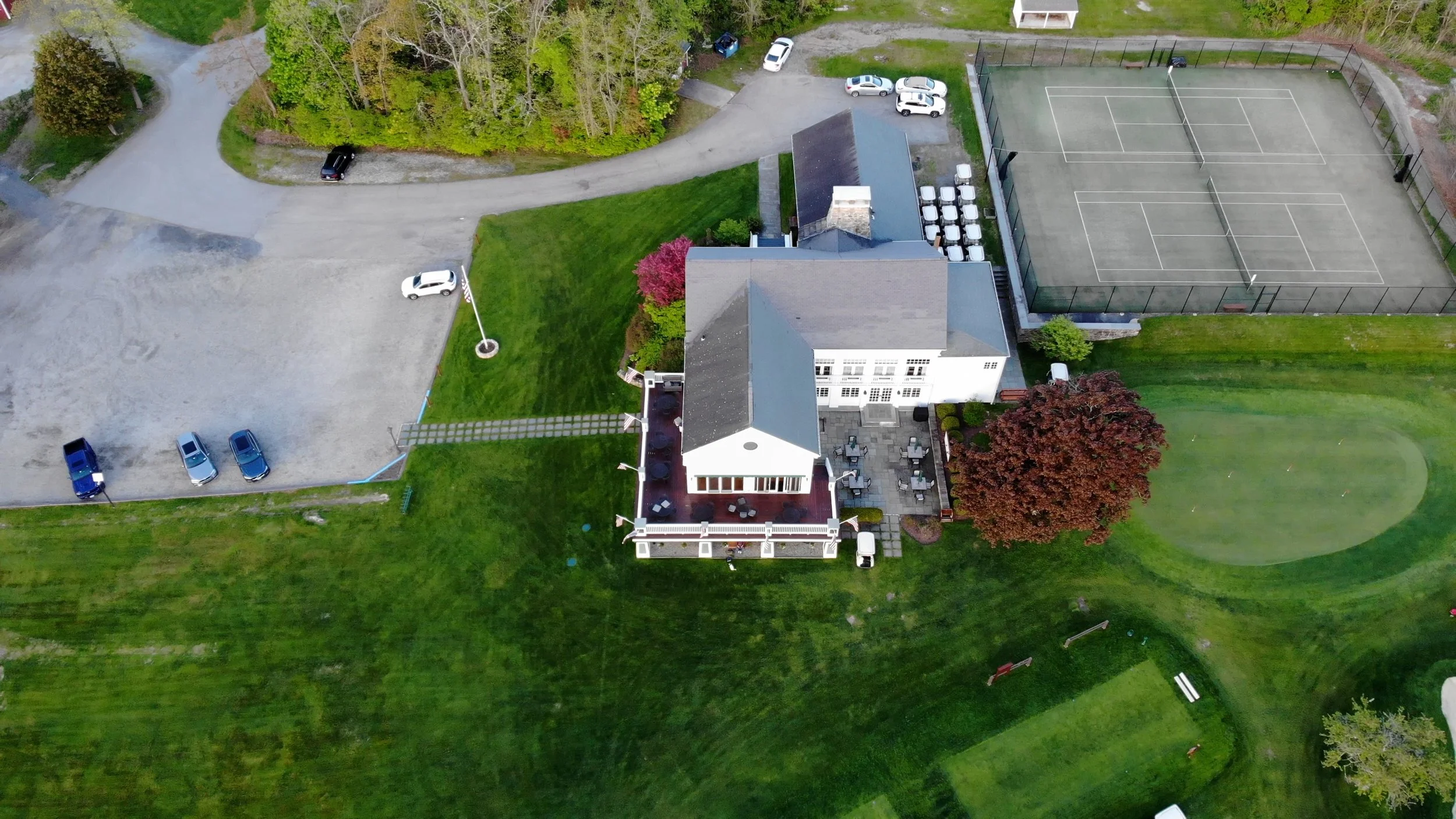 An aerial view of a large house with a patio, surrounded by a well-maintained lawn, trees, and a tennis court with fencing.