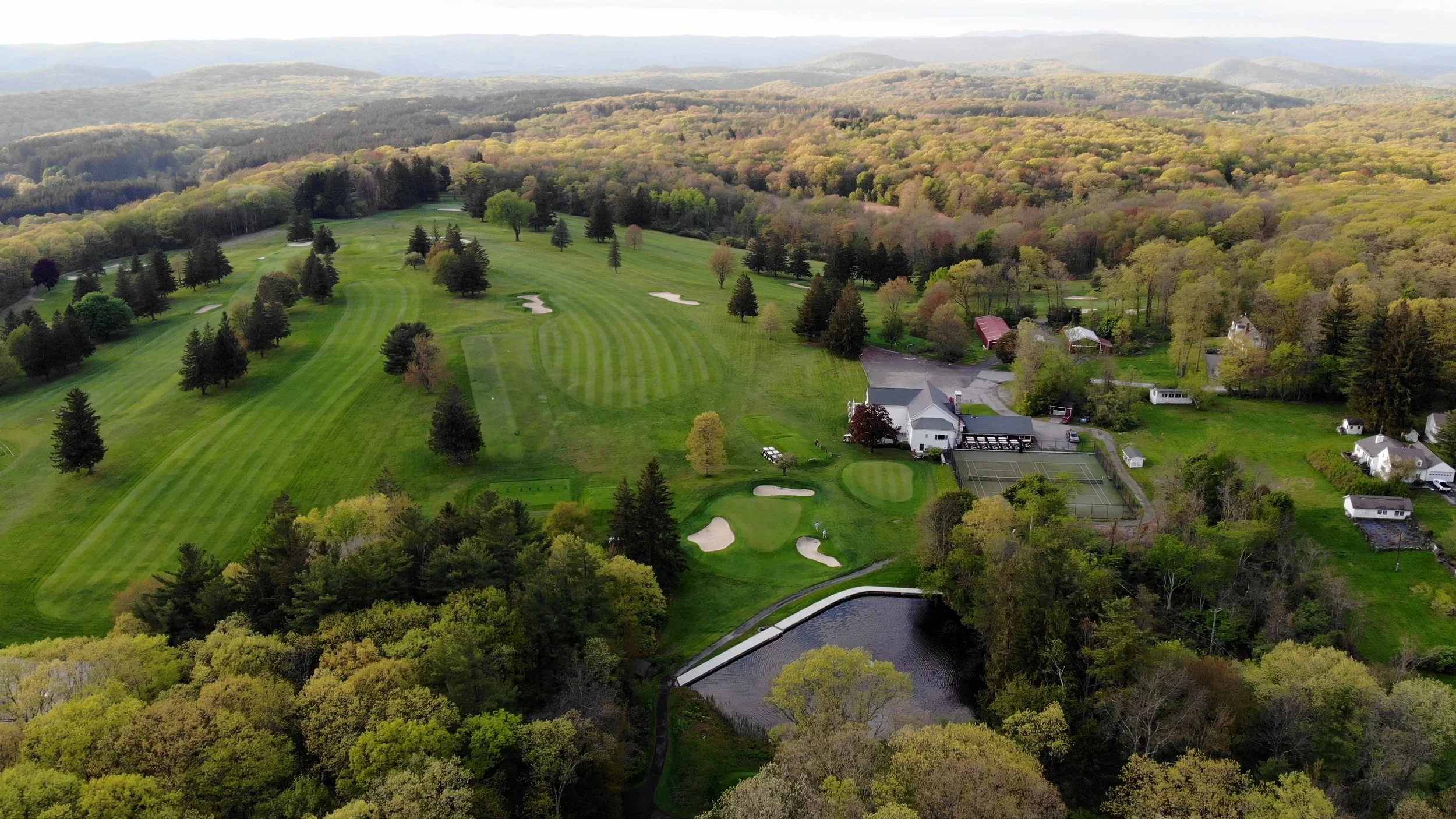 Aerial view of a golf course surrounded by trees and a small community with houses, tennis courts, and a pond in a hilly landscape.