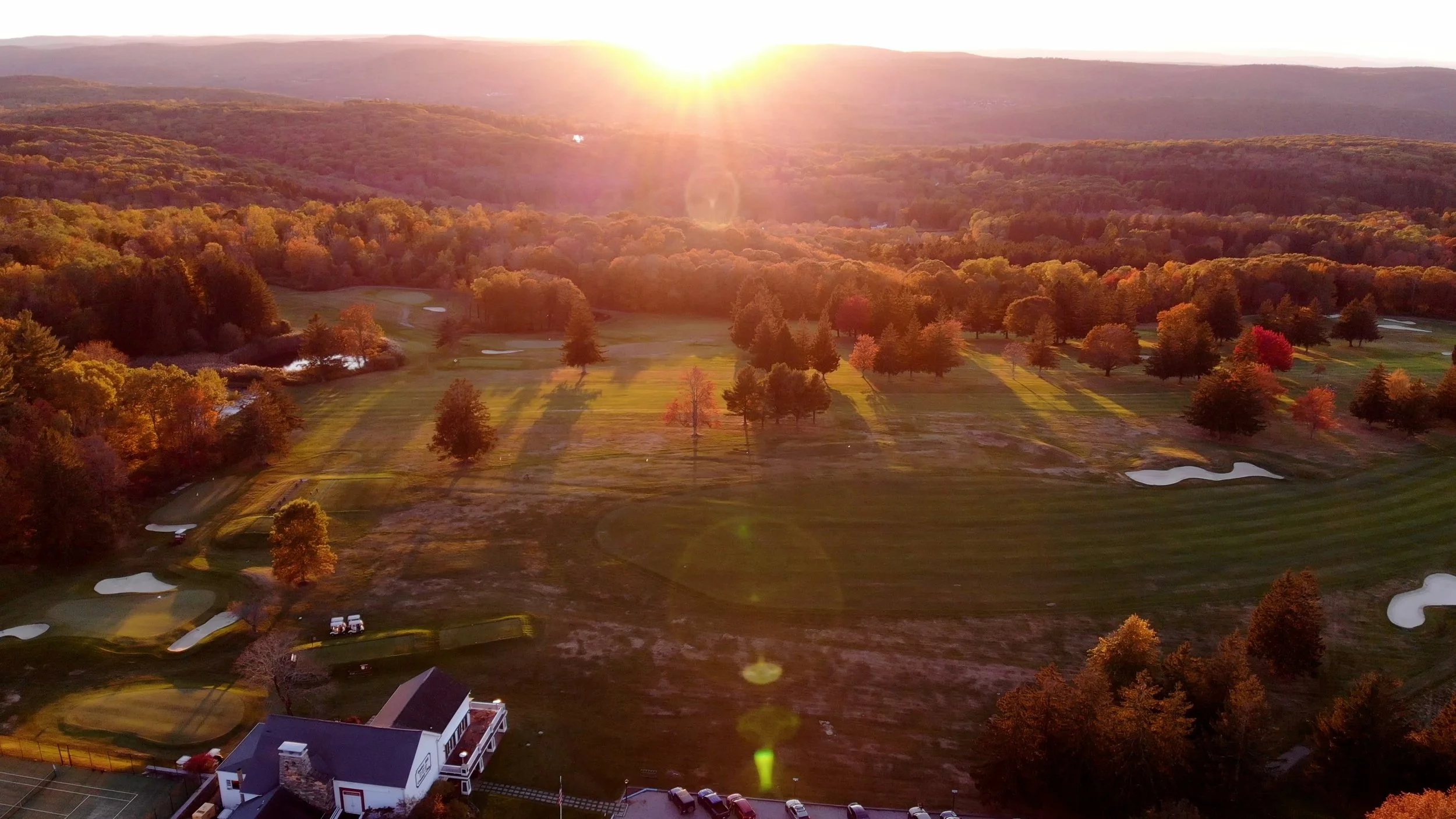 Aerial view of a golf course surrounded by autumn trees with a mountain range in the background, under a setting sun.