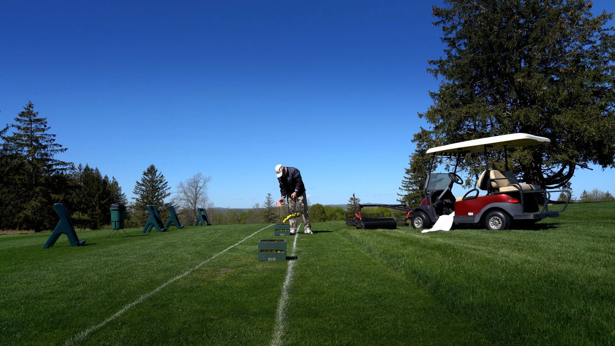 A man on a golf course practicing at a driving range with golf balls, a golf cart, and training equipment under a clear blue sky.