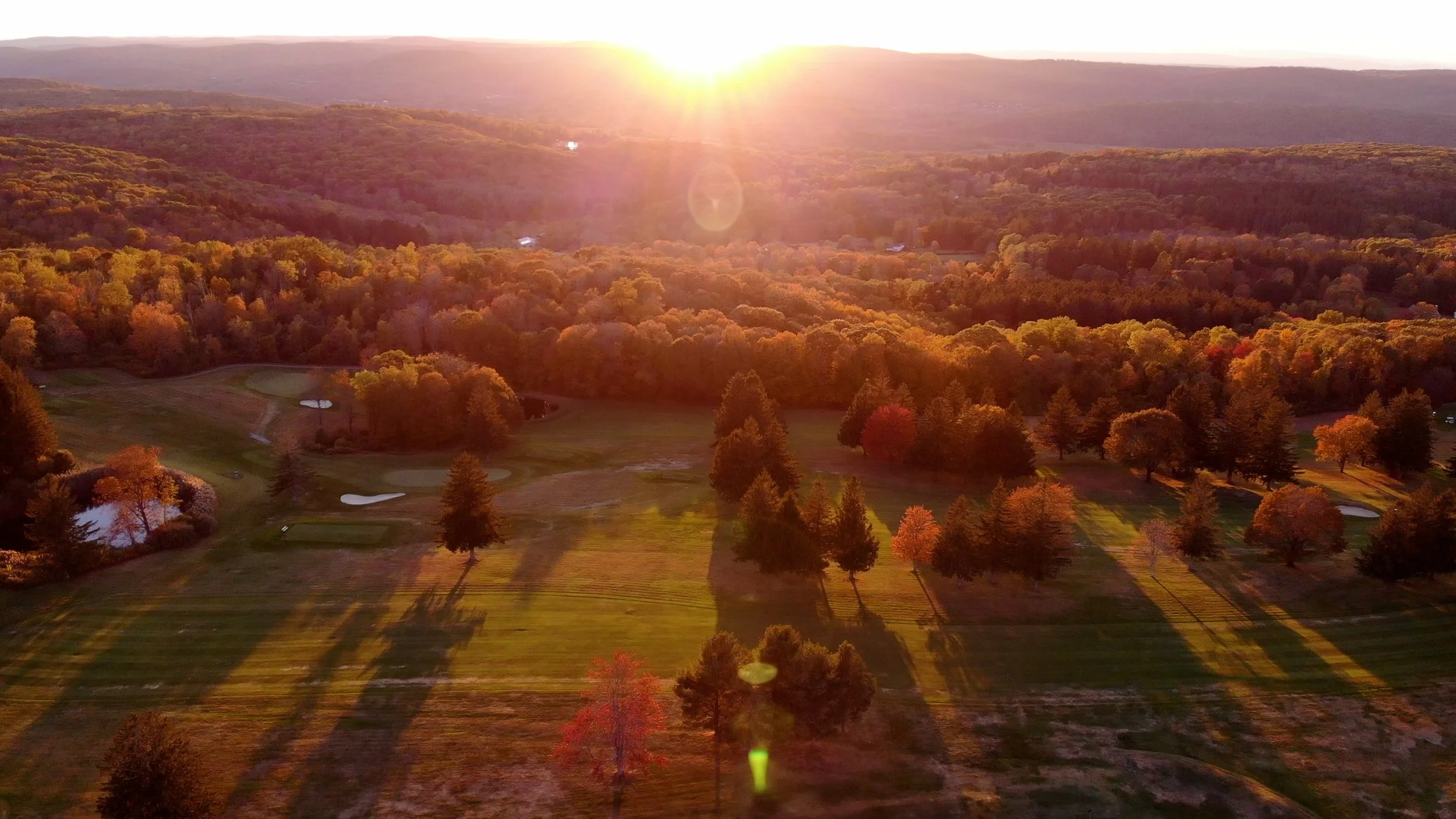 Aerial view of a golf course surrounded by fall trees with a setting sun in the background.