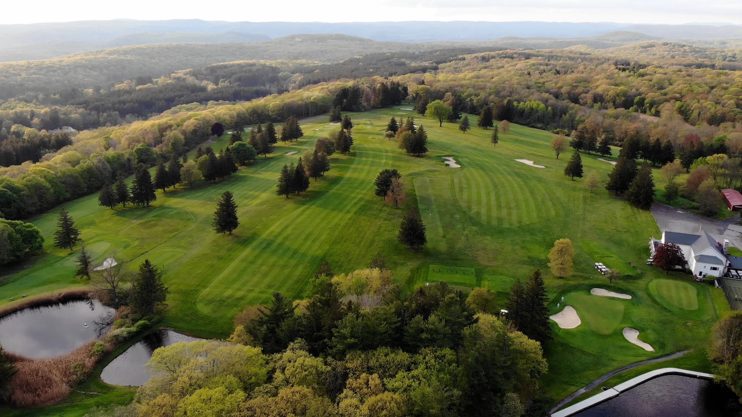 Aerial view of a golf course surrounded by lush green trees and rolling hills, with several sand traps and a pond on the course.