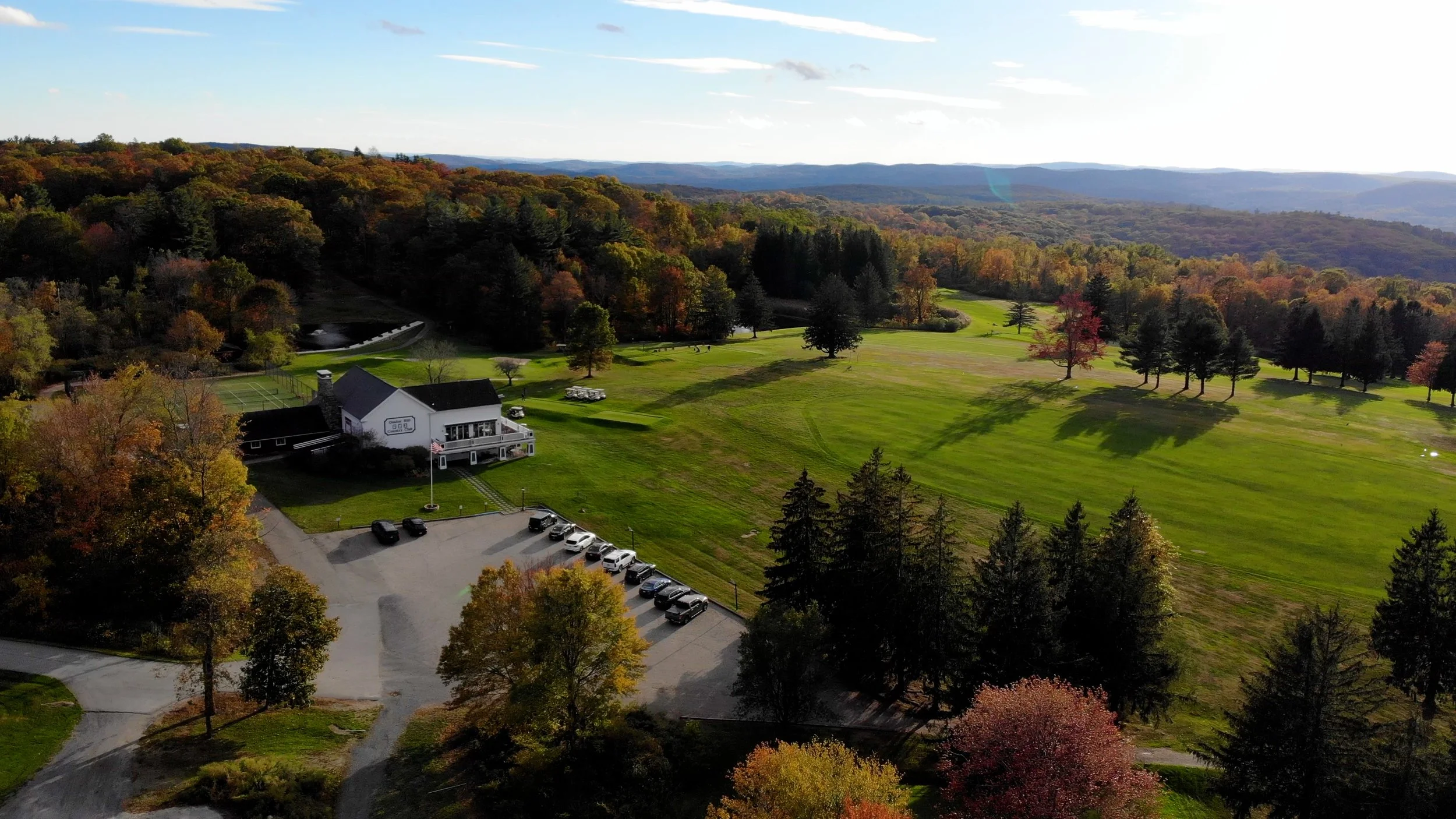 Aerial view of a large grassy area with trees, a building, a parking lot, and distant mountains in the background during fall.