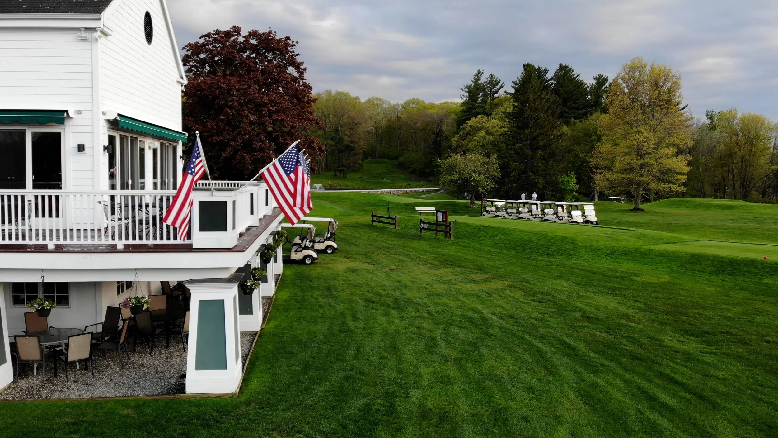 A white golf clubhouse with a deck decorated with American flags, outdoor seating, and a view of a green golf course with multiple golf carts parked in a row. The background features trees with green and red foliage under a partly cloudy sky.