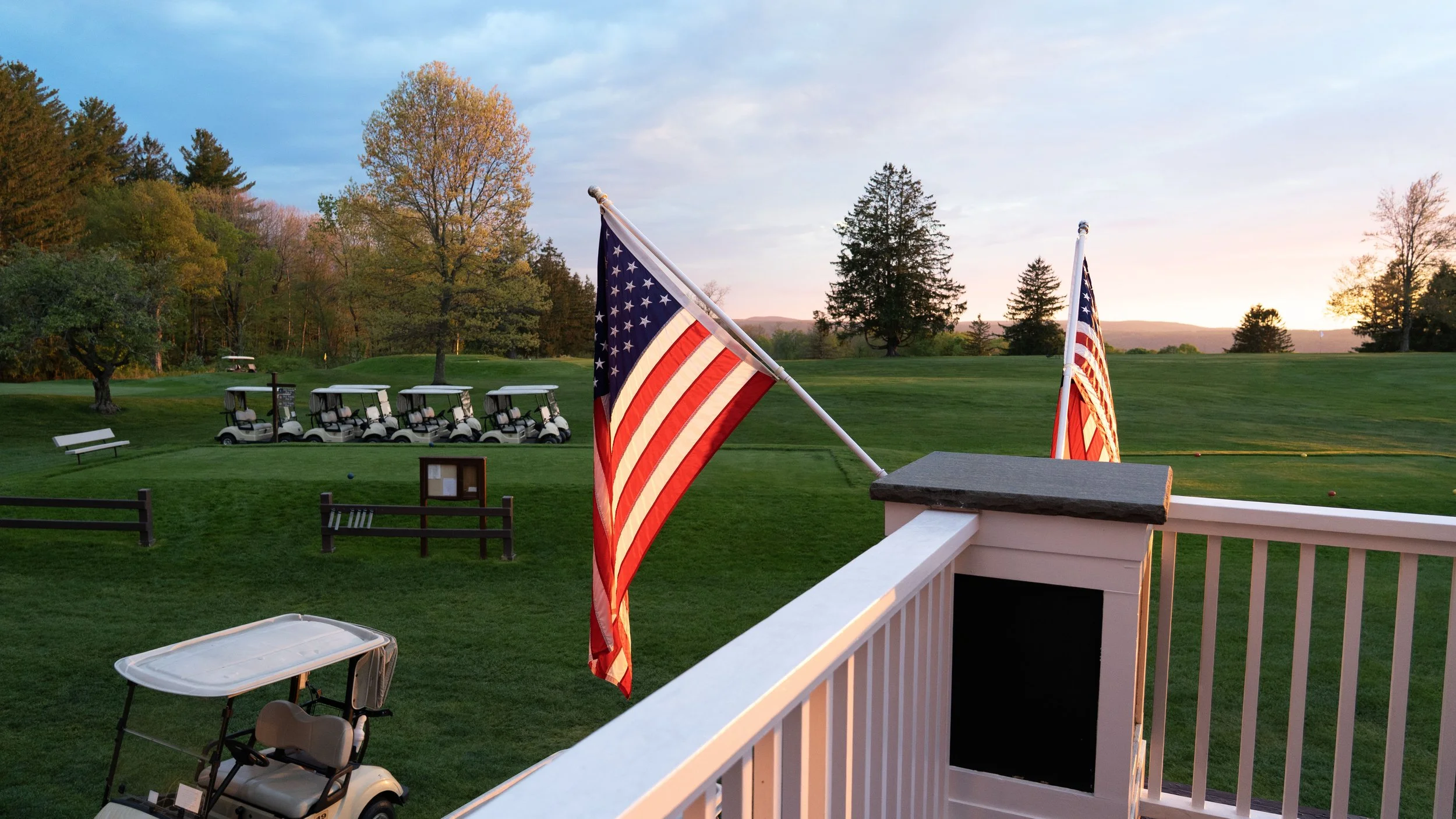 View from a balcony overlooking a golf course with American flags waving, several golf carts parked, and trees with a mountain range in the background during sunset.