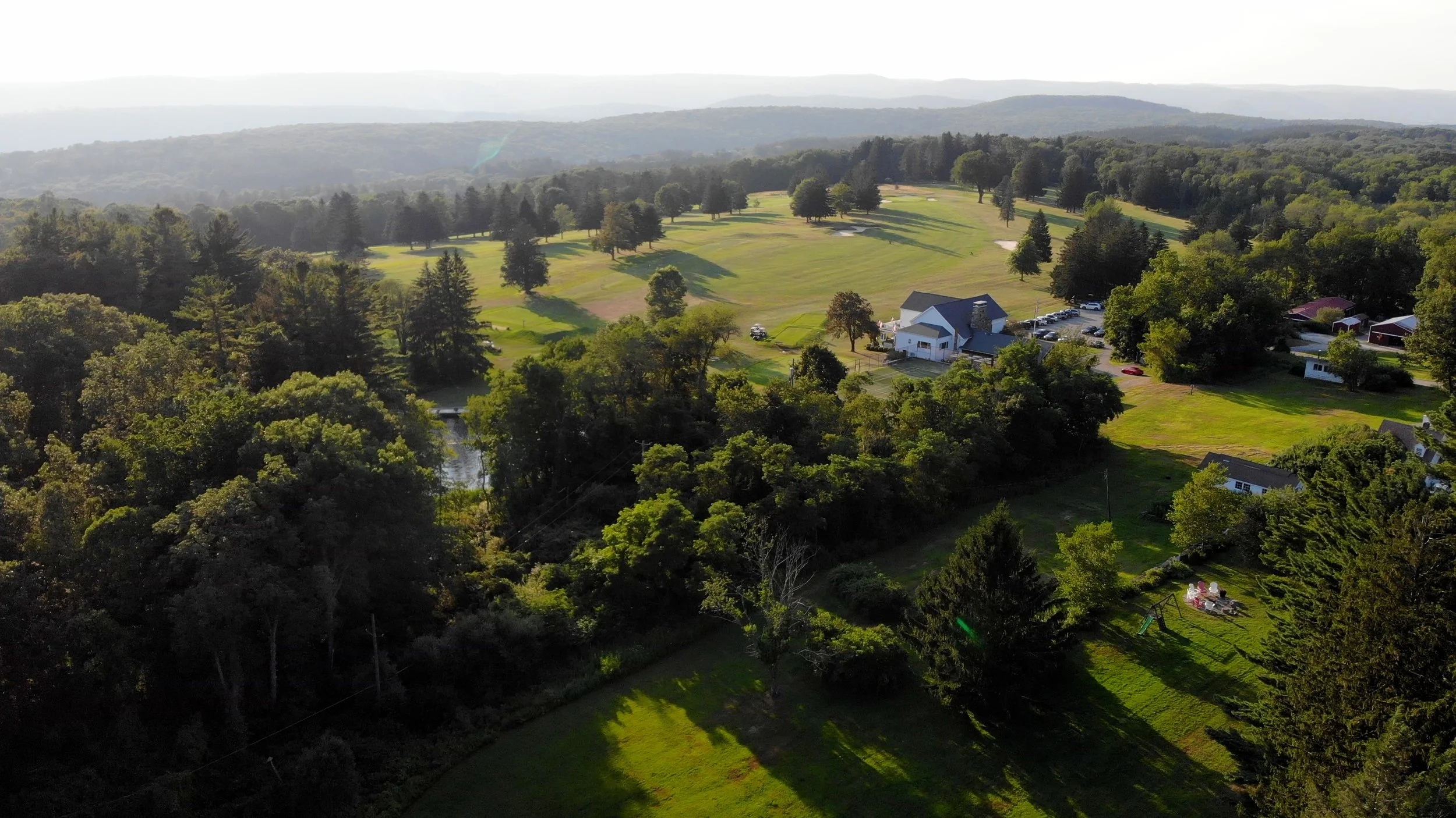 Aerial view of a lush green landscape with a golf course, surrounded by trees and a few houses, with hills in the background.
