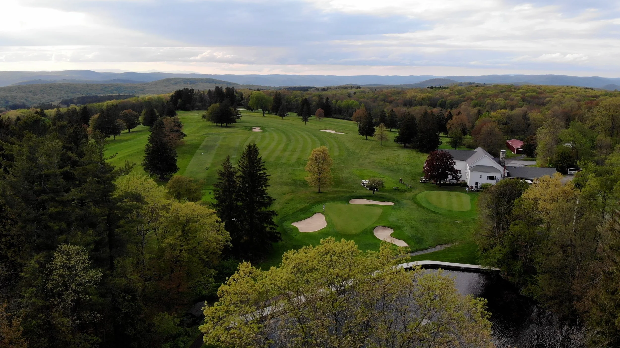 A scenic view of a golf course surrounded by trees and rolling hills with clouds overhead.