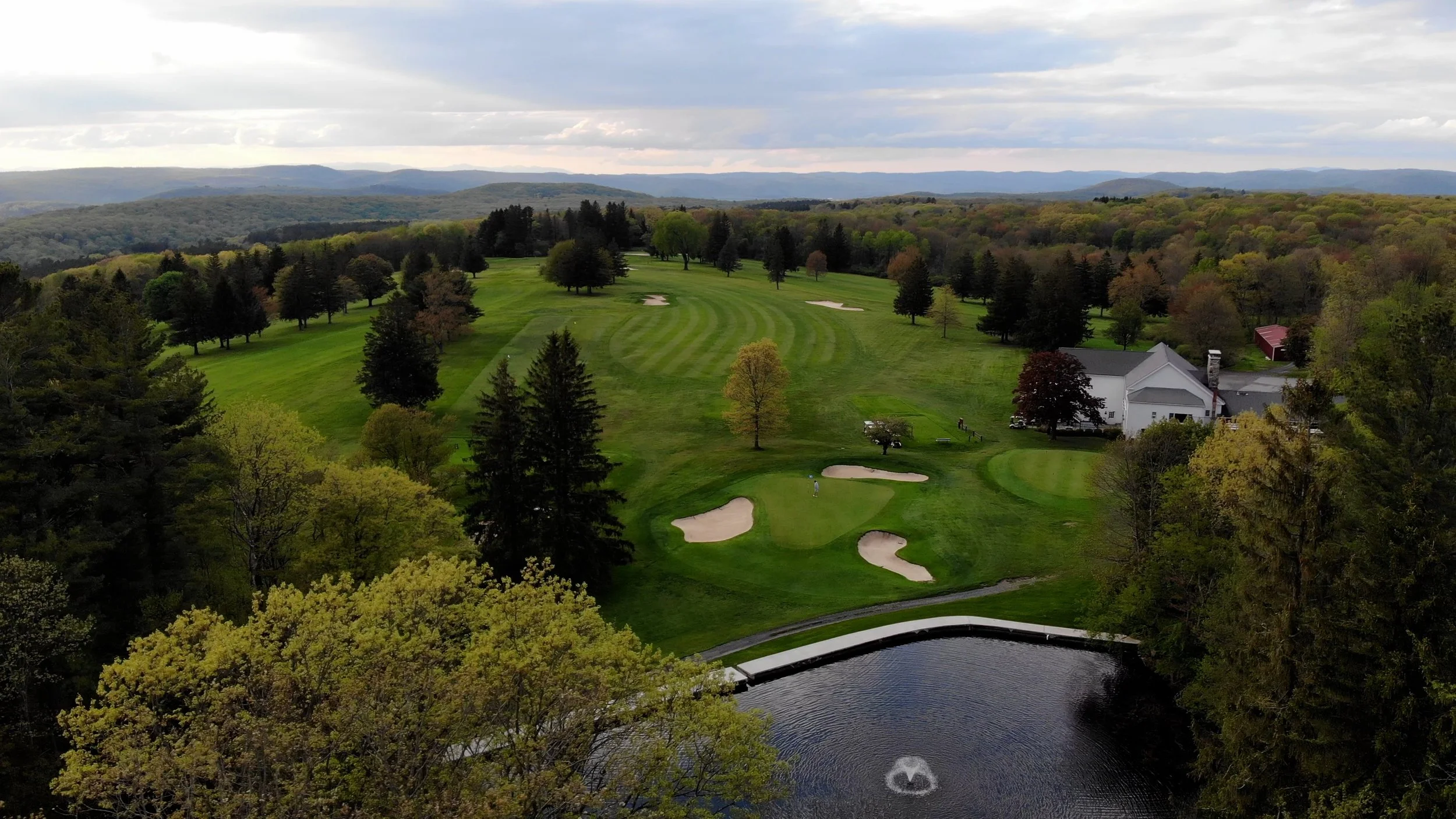 A scenic view of a golf course surrounded by trees, rolling hills, and mountains in the background, with a pond in the foreground and a large white building nearby.