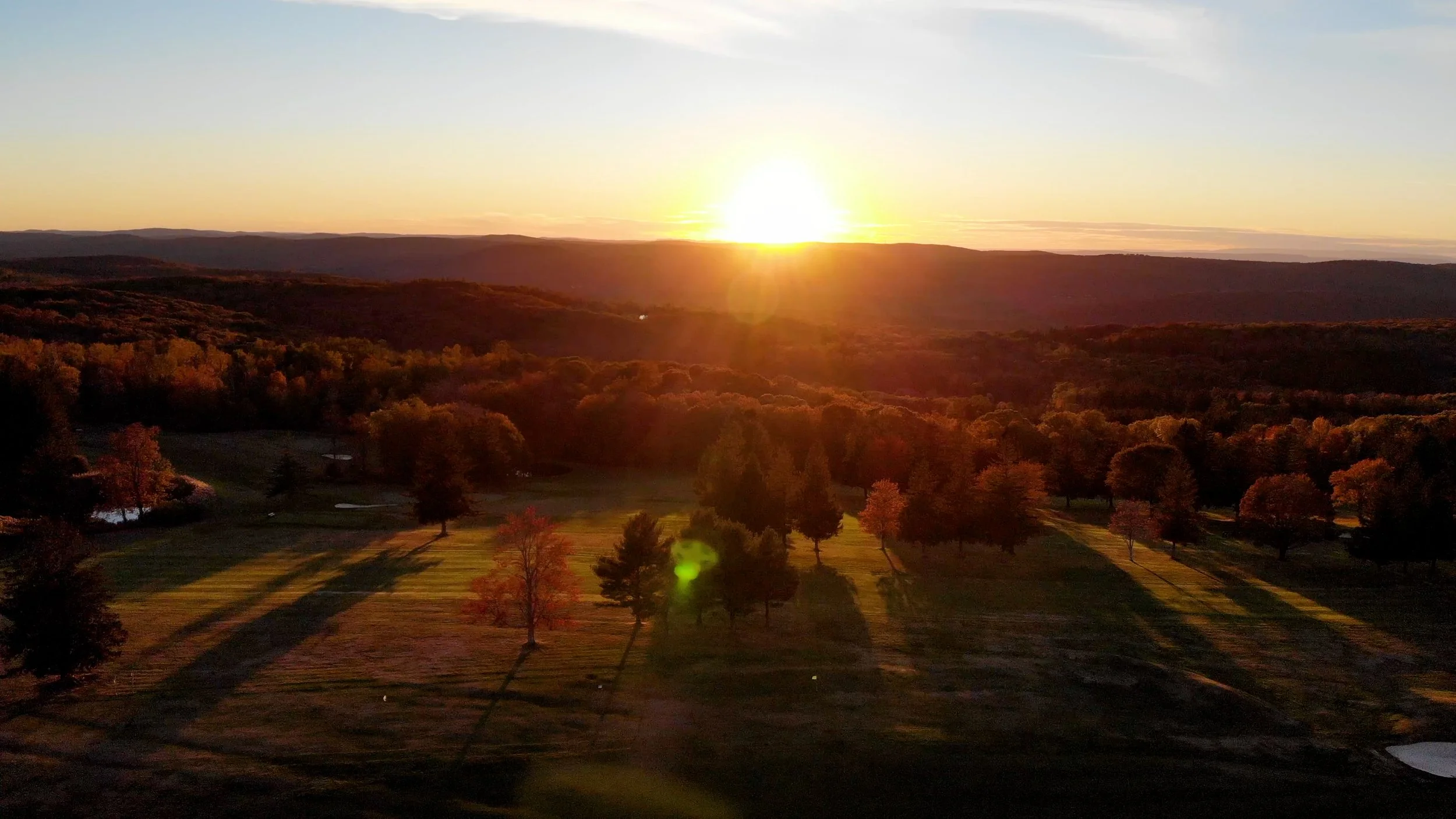A scenic view of a sunset over a hilly landscape with a golf course in the foreground, trees with fall foliage, and mountains in the distance.