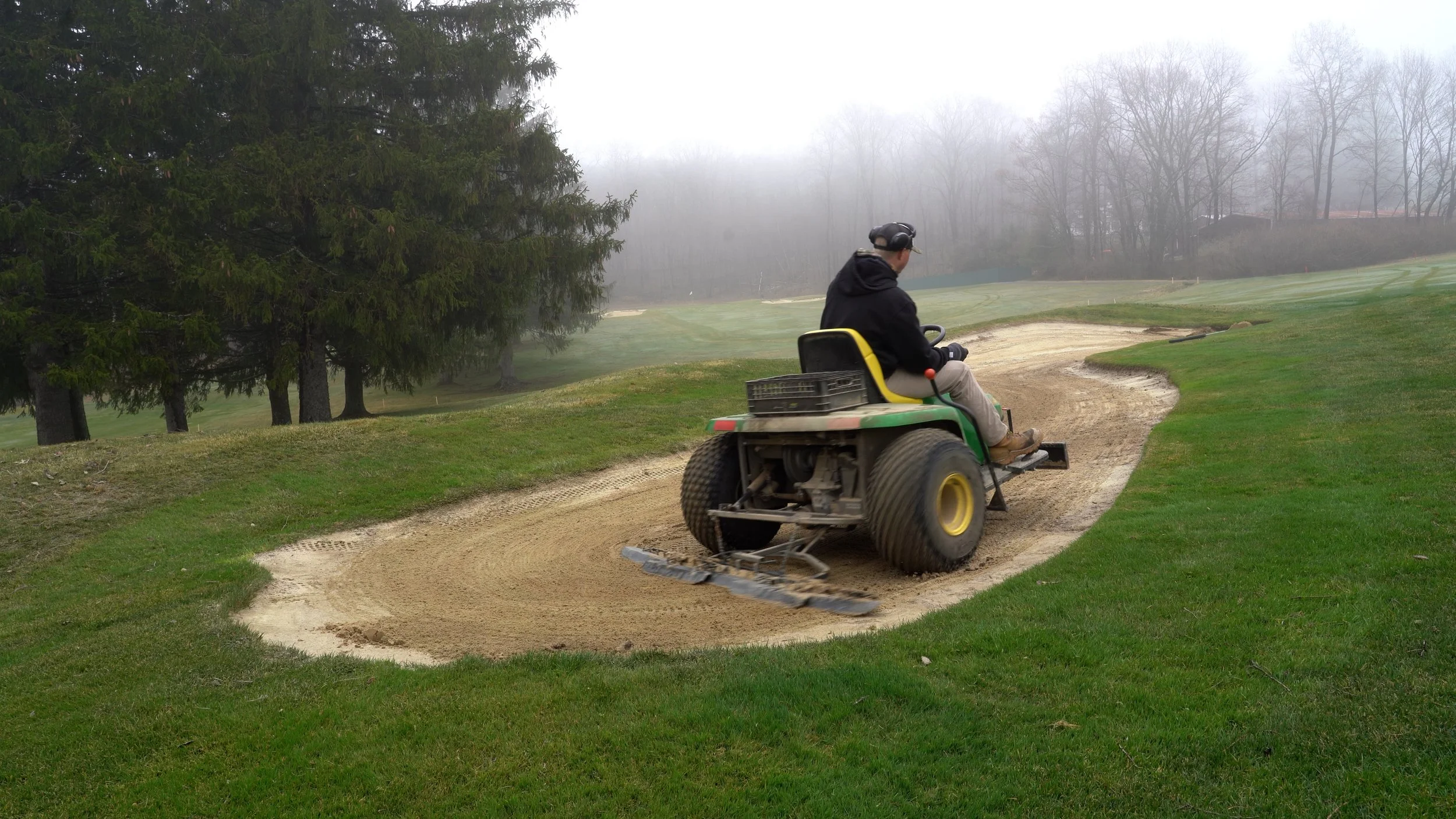 A person in a black hoodie and beige pants riding a greens mower on a sand bunker on a golf course, surrounded by trees and fog.