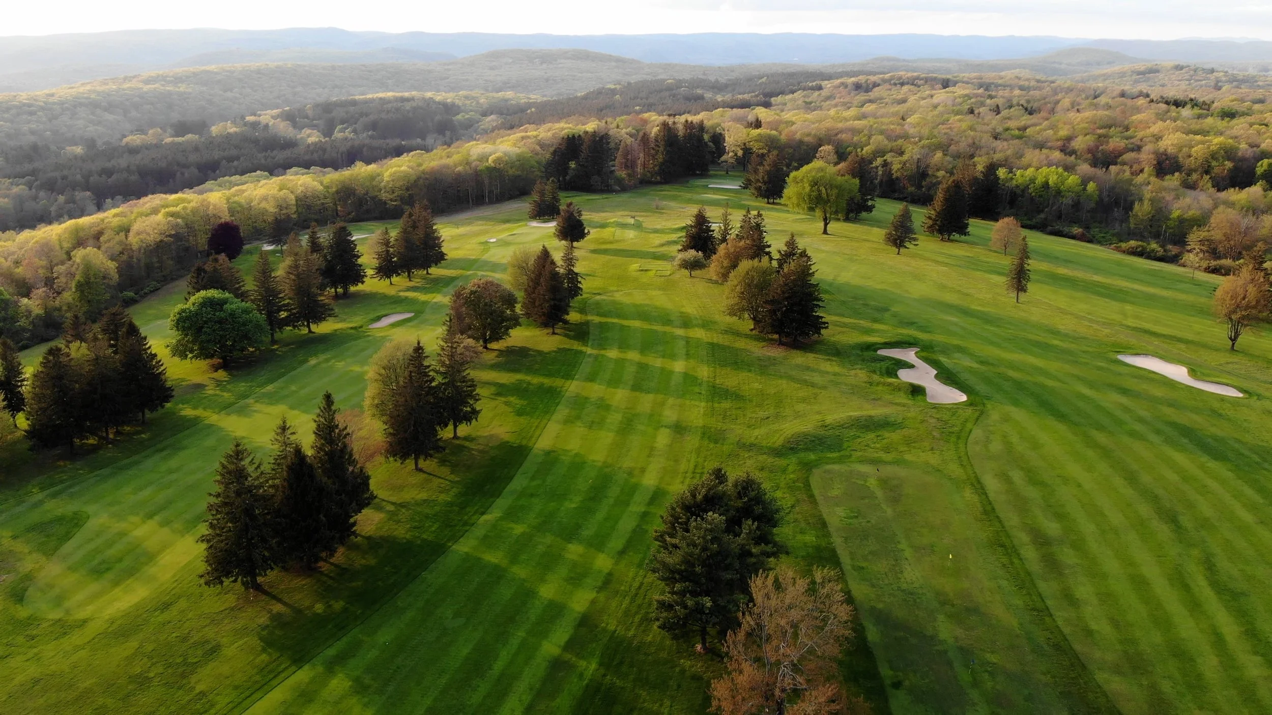 Aerial view of a lush green golf course with sand bunkers, surrounded by numerous trees and rolling hills in the background.