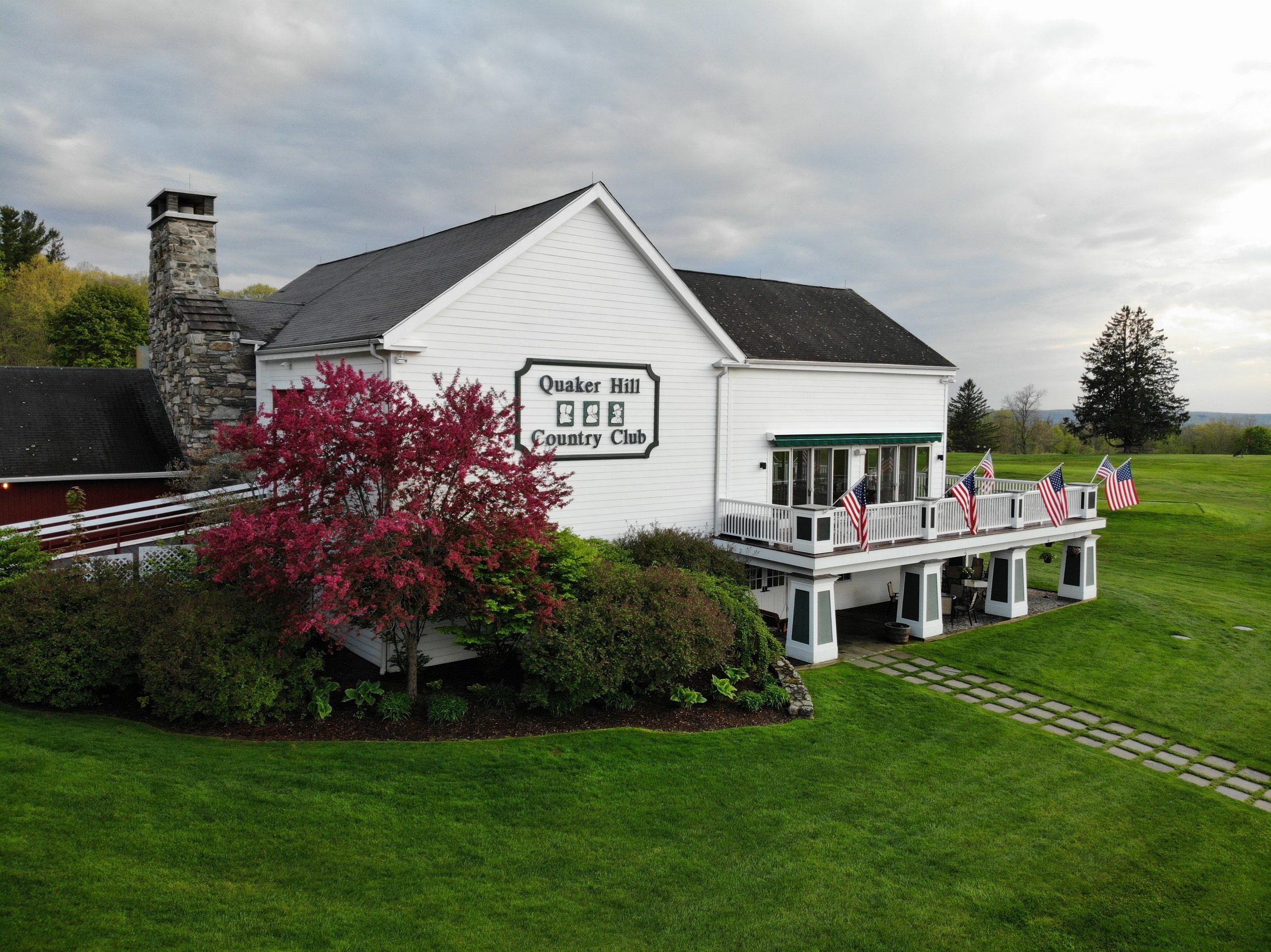 A white building with a sign that reads 'Quaker Hill Country Club,' featuring a deck with multiple American flags. The building has a black roof, a stone chimney, and is surrounded by greenery, including a flowering pink bush in the foreground and tr