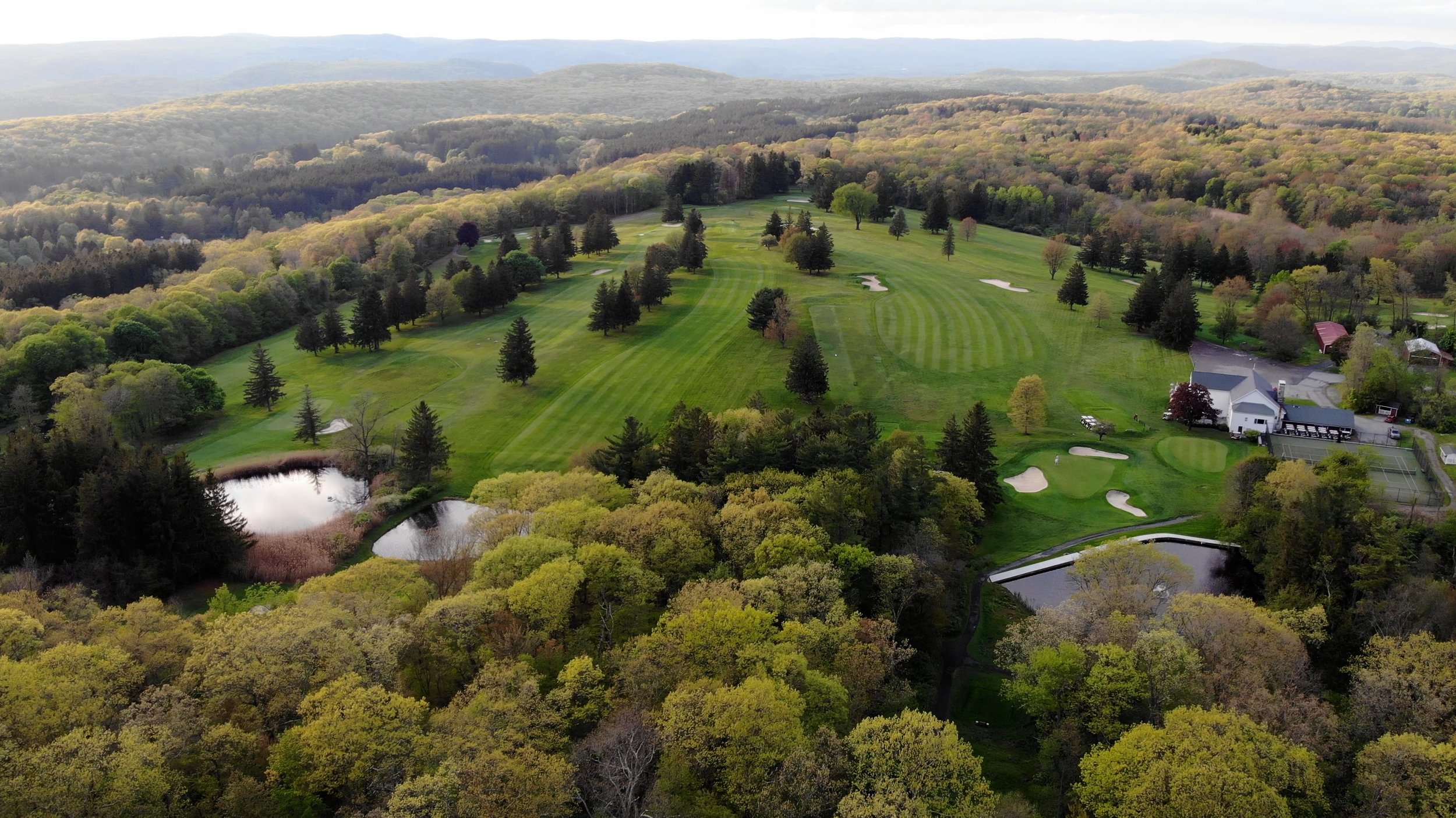 Aerial view of a golf course surrounded by trees on rolling hills with a small pond near the edge of the course.